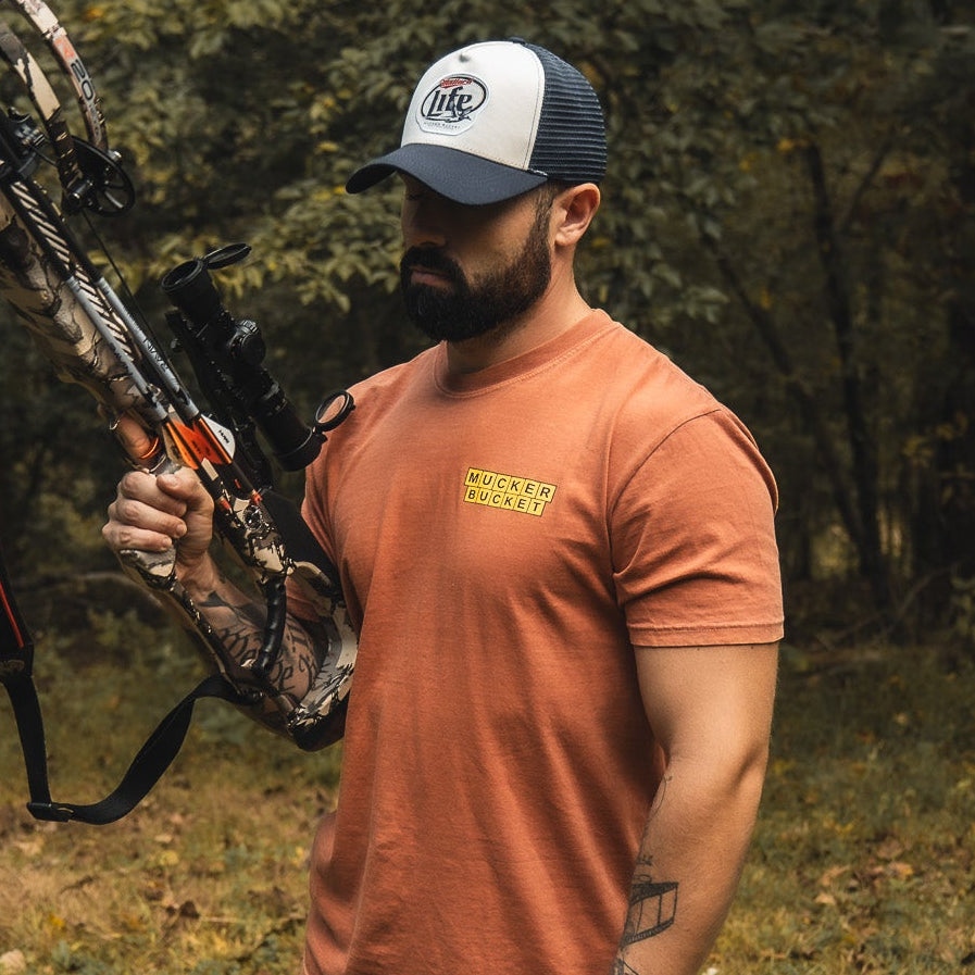 A bearded man wears the Mallard Life Trucker Bucket by My Store, paired with an orange t-shirt, holding a camouflage compound bow outdoors with trees and mallard life in the background.