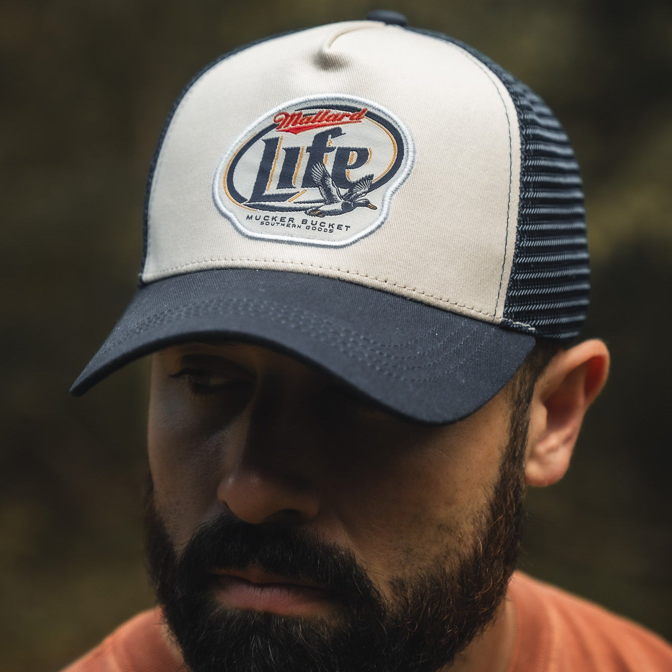 A bearded man wears the My Store Mallard Life Trucker Bucket, a navy and white snapback hat with patch embroidery reading “Mallard Life” and a duck in flight. The blurred outdoor background highlights the cap’s unique look.