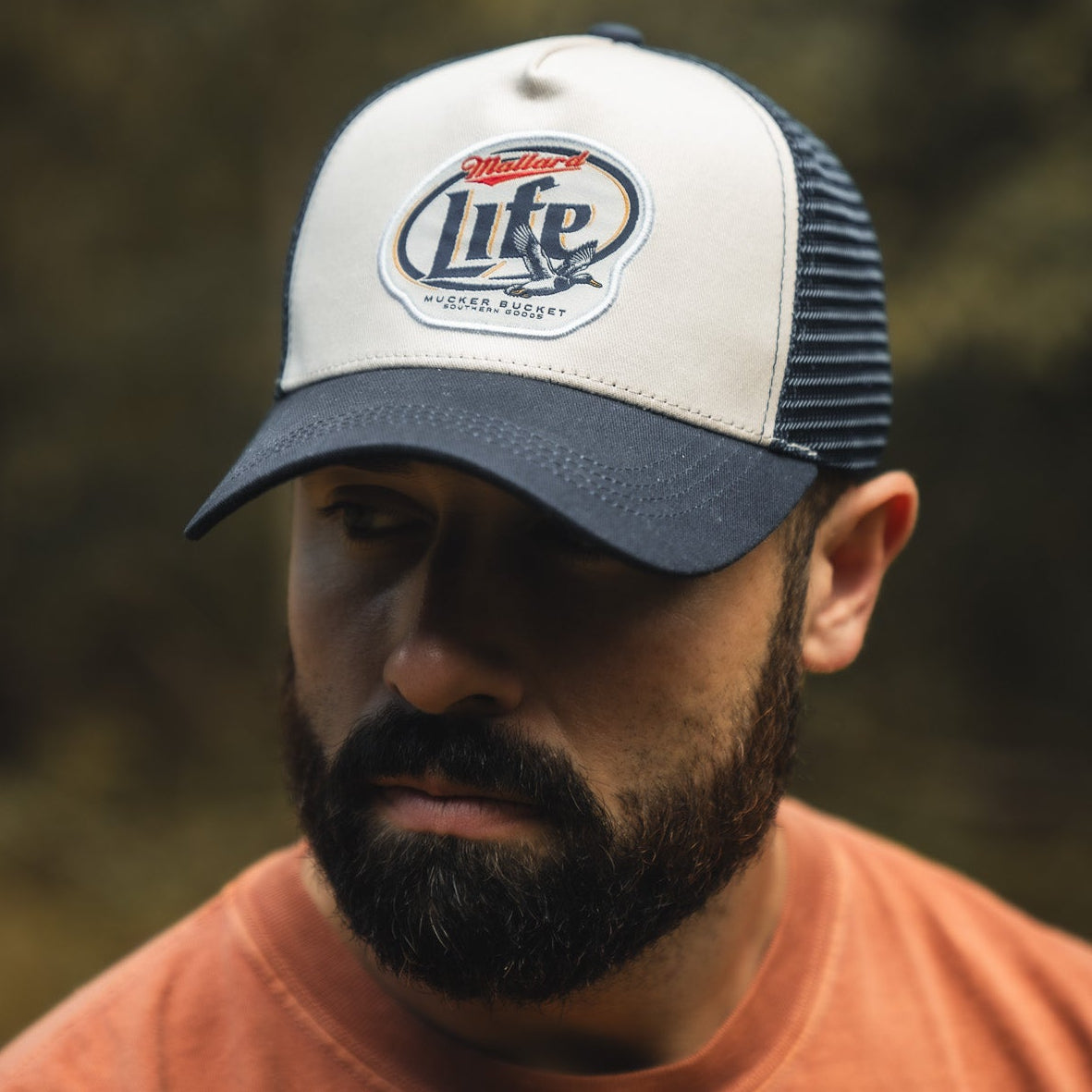 A bearded man in an orange shirt wears a white-and-blue "Mallard Life Trucker Bucket" hat by My Store, featuring patch embroidery, and looks down with a neutral expression against a blurred outdoor background.