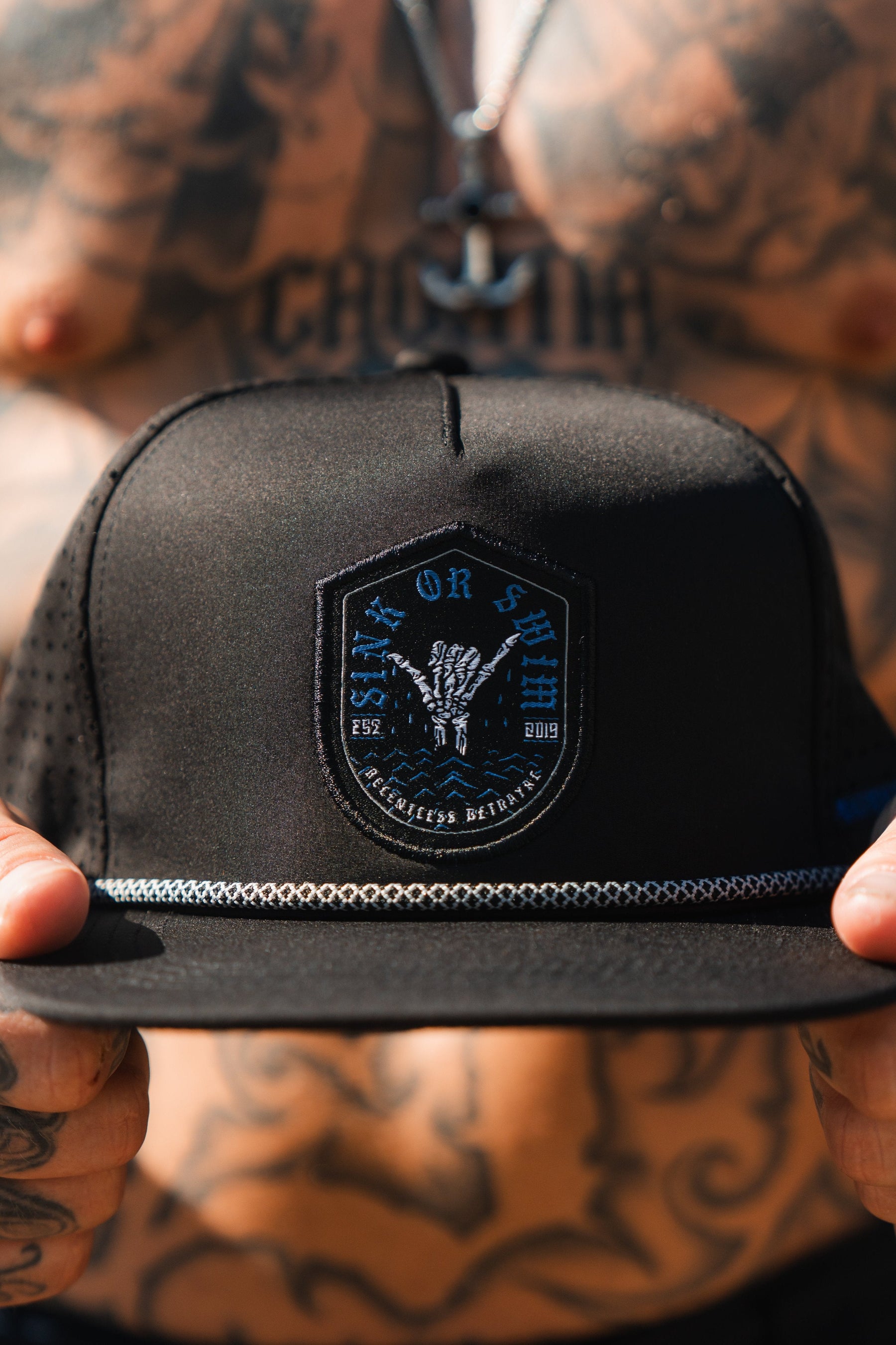 A tattooed person dons the Sink Or Swim Snapback by Relentless Betrayal. This black cap, featuring a gothic skeletal hand emblem and chain-like brim, symbolizes resilience and the journey of overcoming challenges.