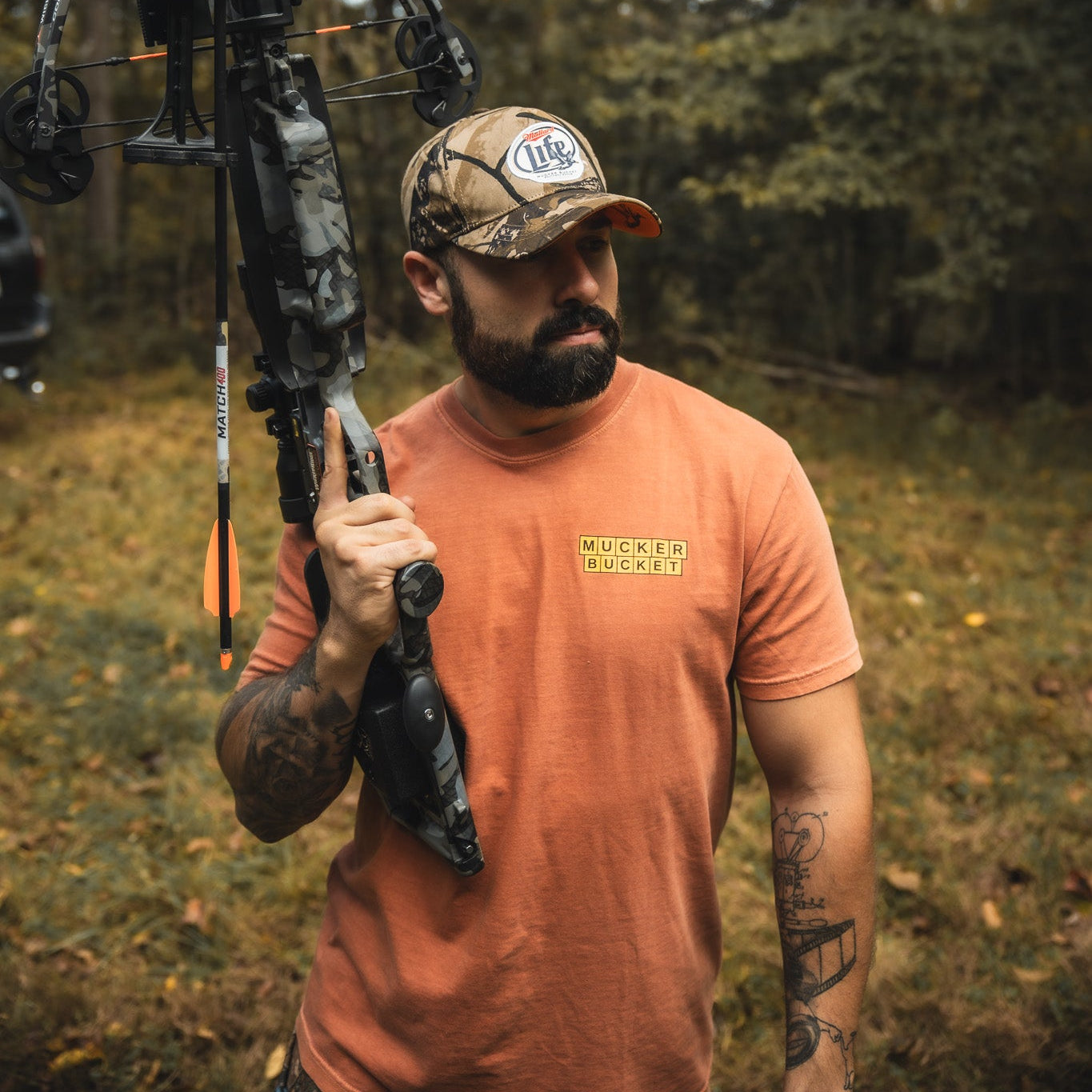 A bearded man stands outdoors holding a compound bow over his shoulder. He wears a Mallard Life Deadwood Camo Bucket from My Store. Trees and greenery fill the background.