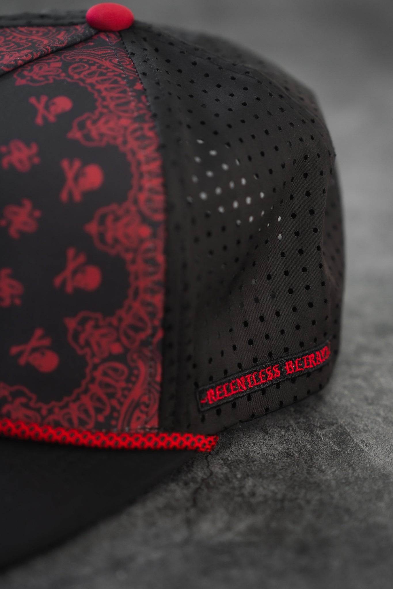 Close-up of a black mesh All Gas No Brakes Premium Snapback by Relentless Betrayal, showcasing red embroidery with a skull and crossbones amid ornate patterns. The cap, echoing a bandana print hat, has RELENTLESS BE3ARD on the side and rests on a gray textured surface.