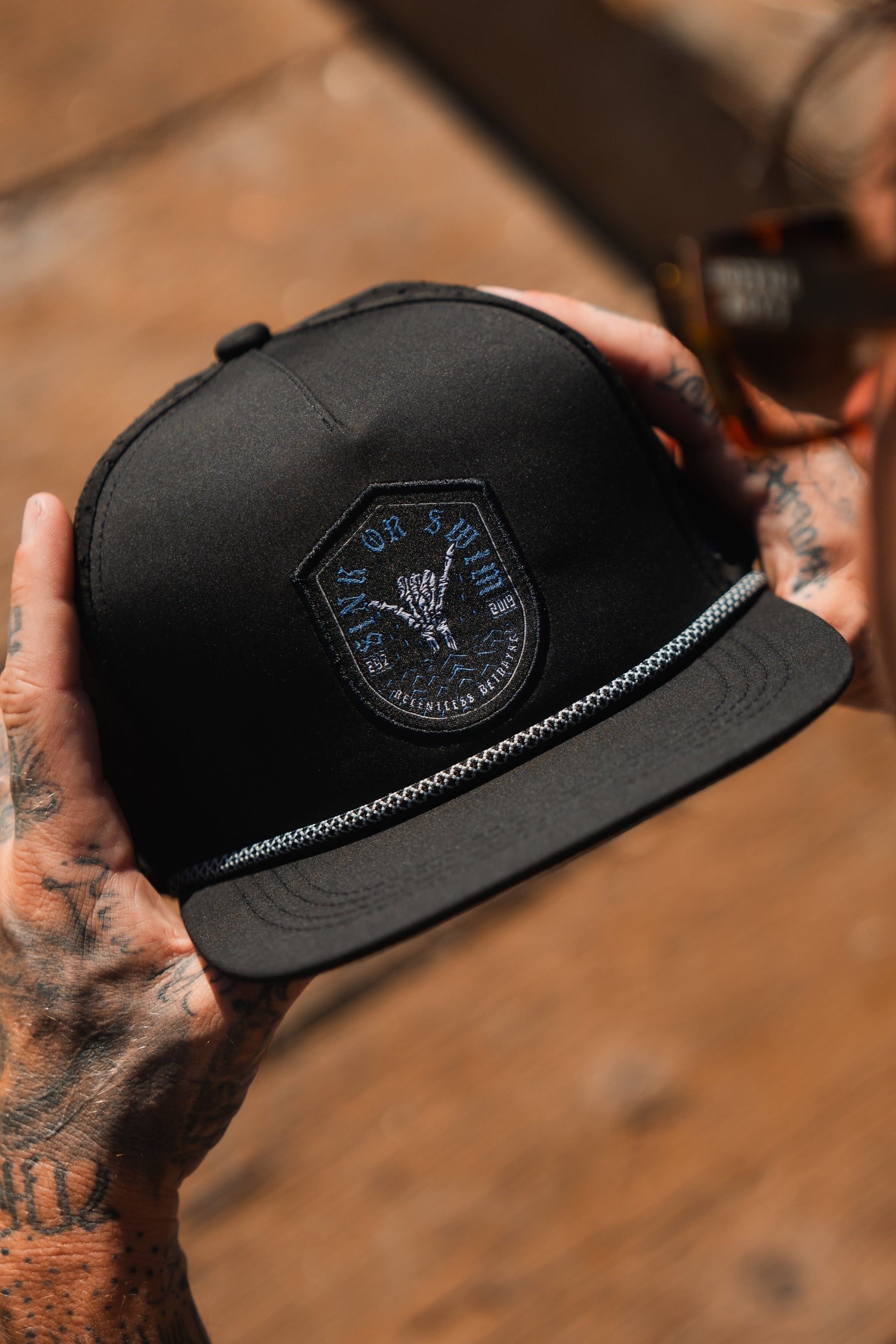 Tattooed hands, symbols of resilience, hold the Sink Or Swim Snapback by Relentless Betrayal, a black cap with a skeletal hand patch and chain-detailed brim. Sunglasses symbolizing overcoming challenges peek from the background.