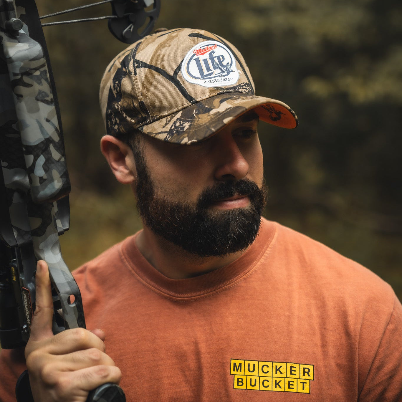 A bearded man in an orange shirt holds the Mallard Life Deadwood Camo Bucket by My Store, wearing a camo cap and looking right with a blurred outdoor background.