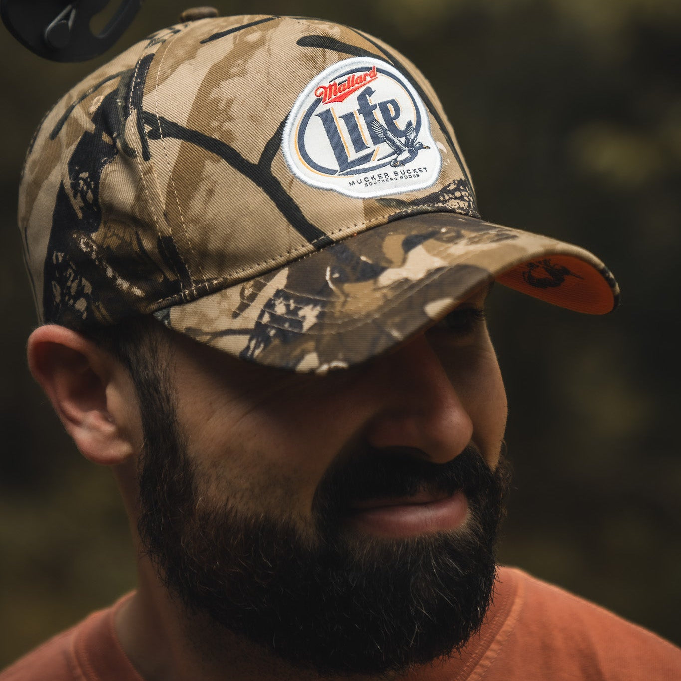 A bearded man stands outdoors wearing a Mallard Life Deadwood Camo Bucket hat from My Store and a burnt orange shirt, with a blurred leafy background.