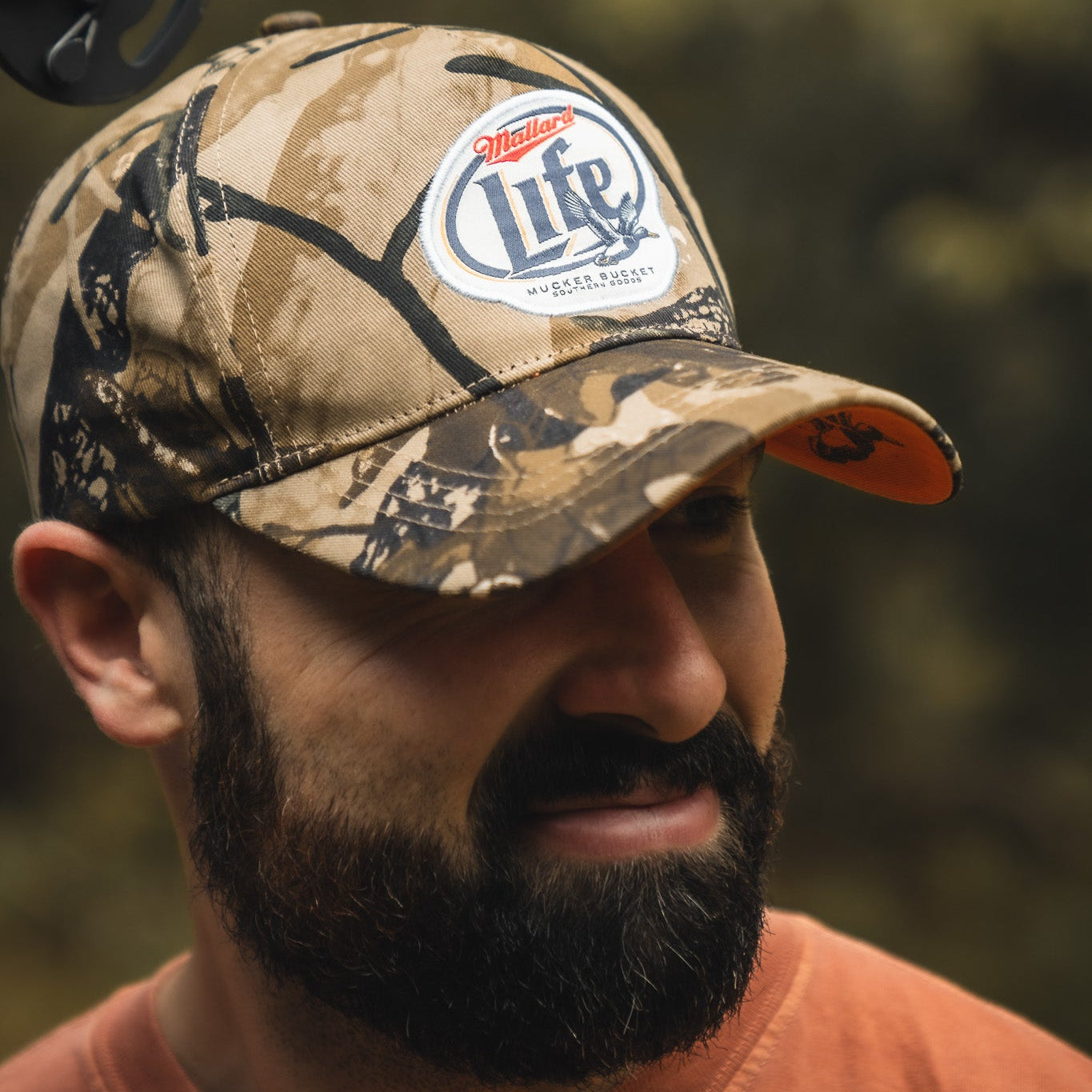 A bearded man stands outdoors wearing the Mallard Life Deadwood Camo Bucket by My Store and an orange shirt. The background is blurred with earthy tones.