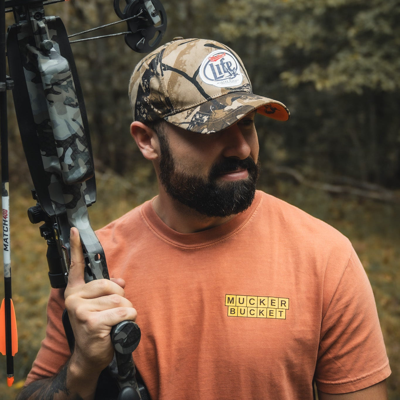 A bearded man in an orange t-shirt and camo hat holds the My Store Mallard Life Deadwood Camo Bucket over his shoulder, standing outdoors in a wooded area.