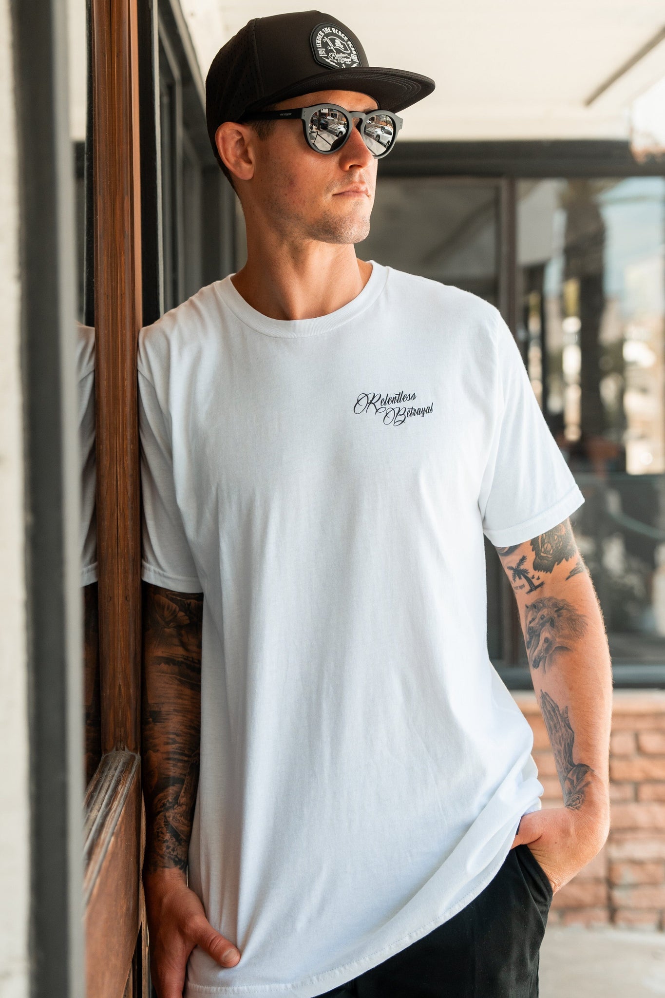 Leaning against a wall, a tattooed individual wearing Relentless Betrayals Skilled Sailor White T-Shirt sports sunglasses and a black cap. One hand rests in their pocket as gleaming glass windows of a building reflect in the background.