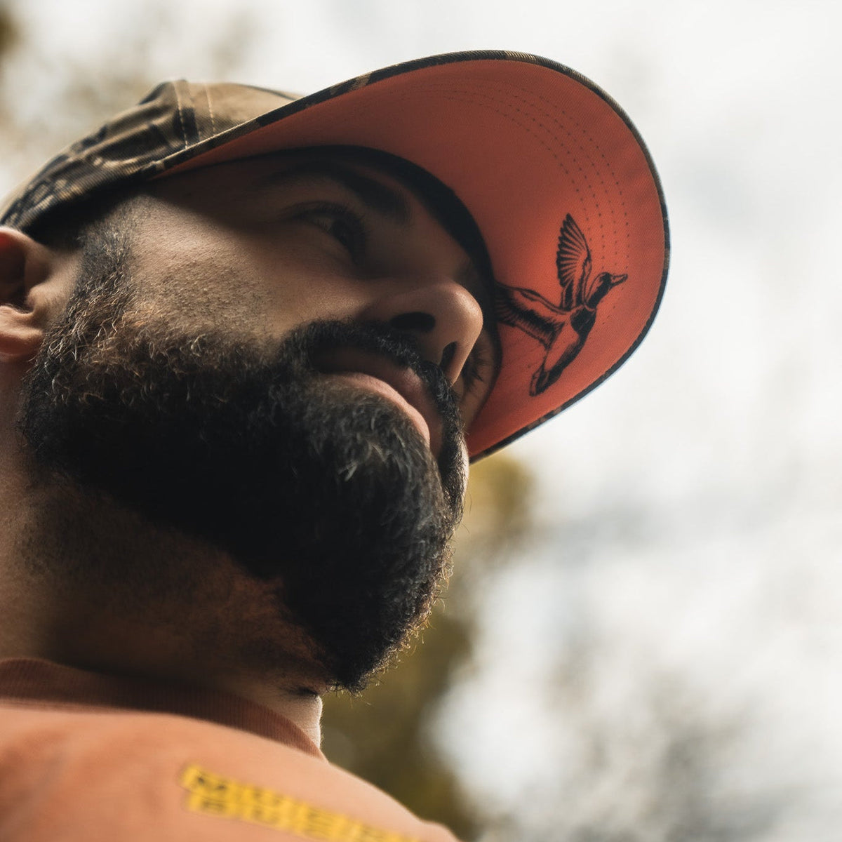 A bearded person wears the Mallard Life Deadwood Camo Bucket by My Store, looking upward against a blurred outdoor background. The low-angle photo highlights the bucket hat’s unique camo style.