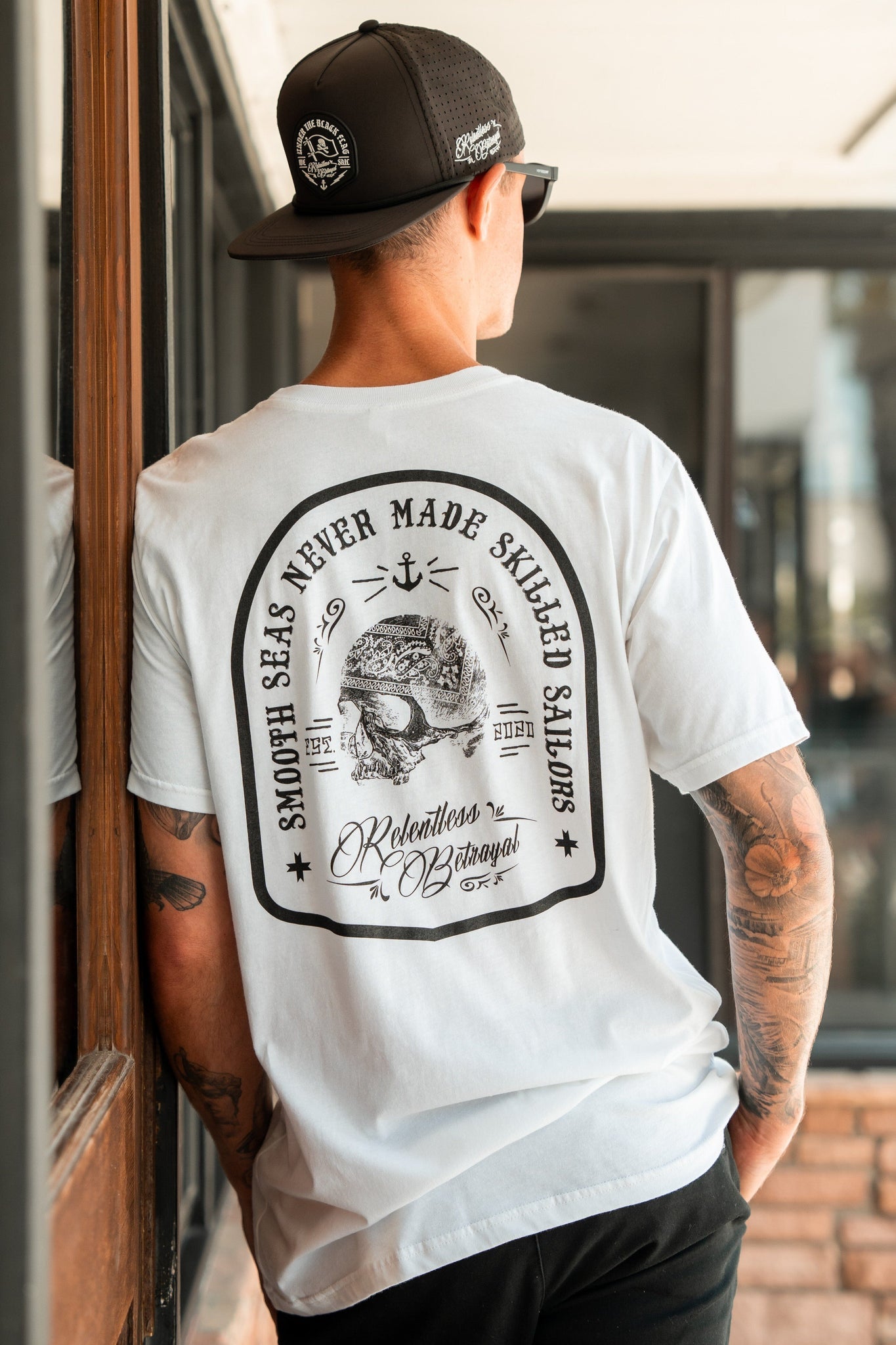 Wearing a Skilled Sailor White T-Shirt by Relentless Betrayal with Smooth seas never made skilled sailors, and a black cap, a man stands by the window, embodying resilience.