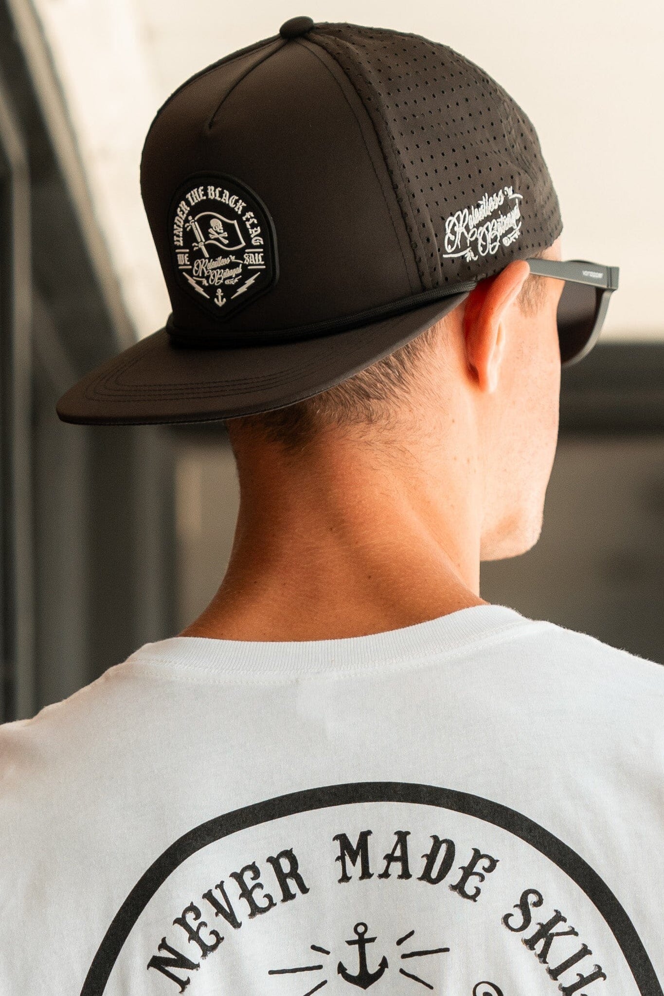 Wearing a Black Flag Snapback- Black by Relentless Betrayal, featuring an anchor logo, and a matching white T-shirt with NEVER MADE SKILLS, RESPECT, the individual in sunglasses effortlessly exudes a pirates life vibe as they face away from the camera.