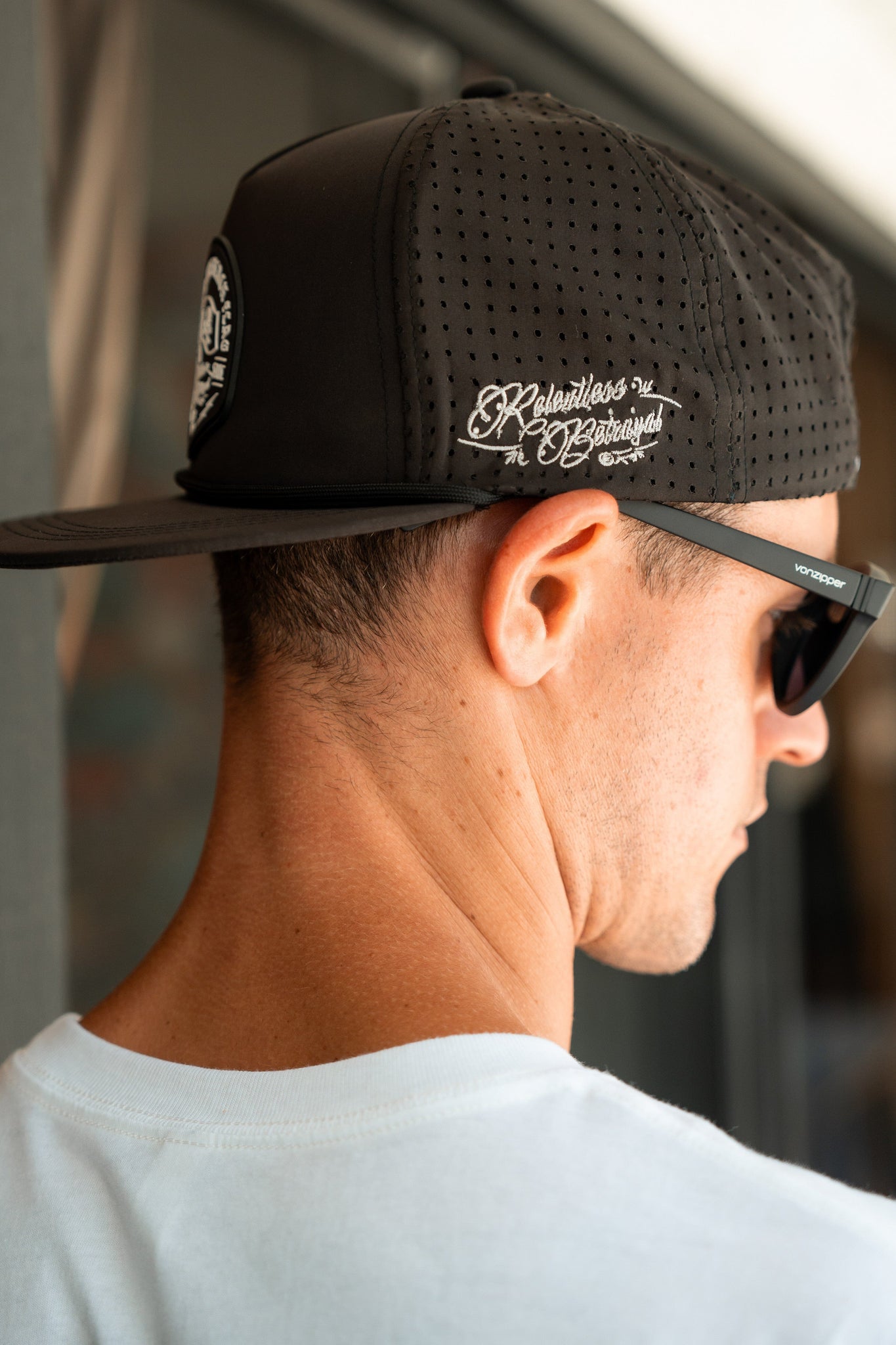 Someone wearing a Relentless Betrayal Black Flag Snapback cap backward over a white shirt, styled with black sunglasses. The black cap features decorative white text and a snapback enclosure, set against a blurred background.