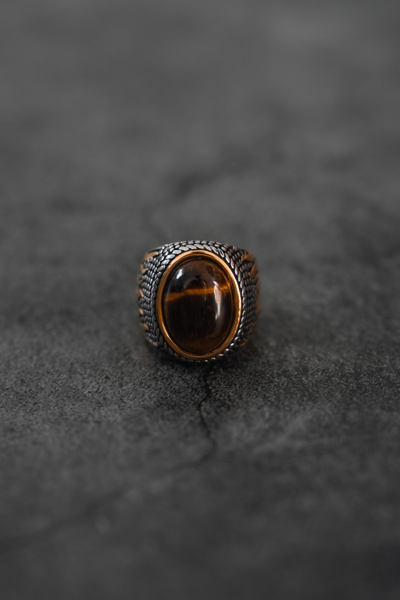The Relentless Betrayal Inferno Ring showcases a large brown oval gemstone set in an ornate, textured metal band, elegantly resting on a dark surface.