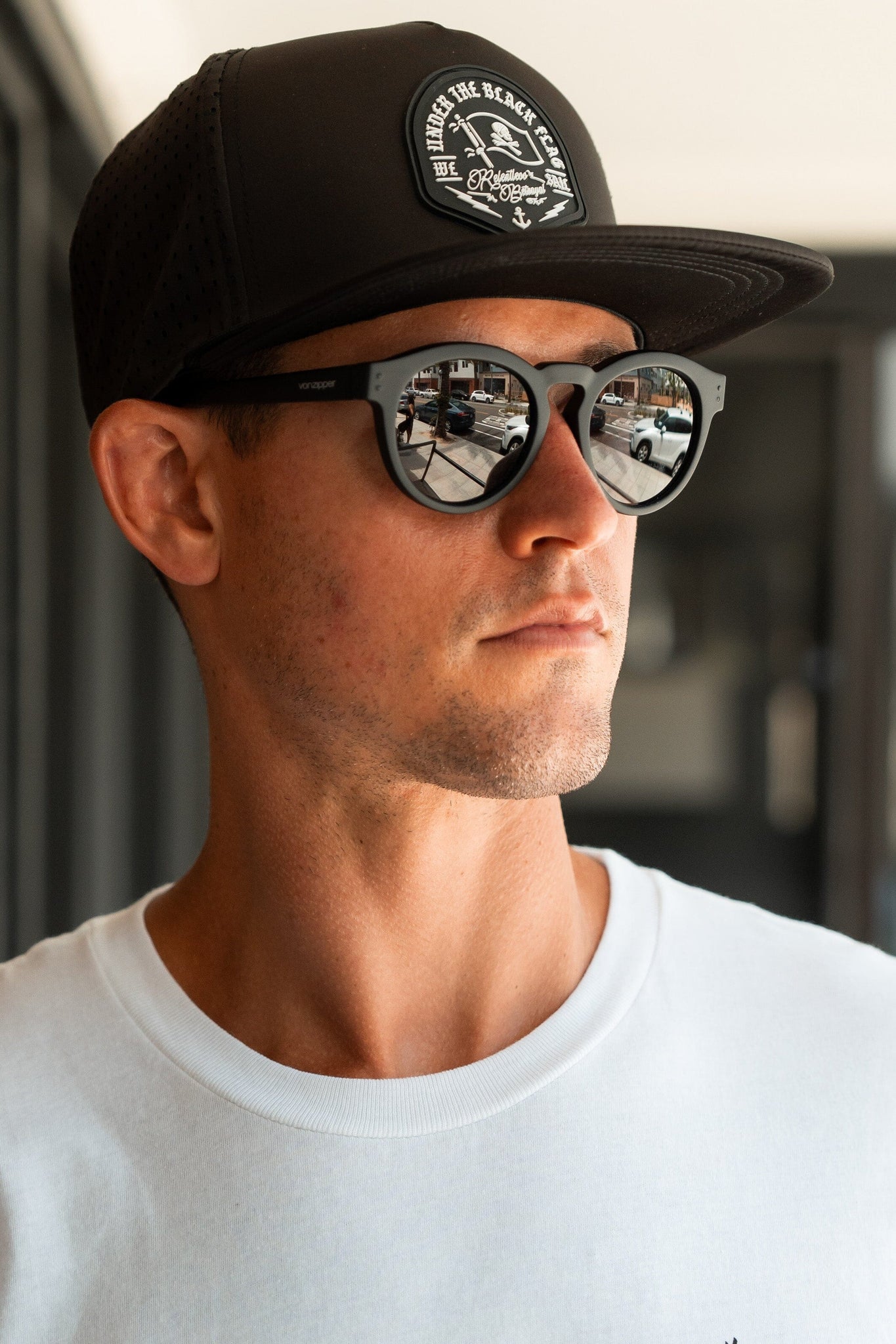 A man wearing the Black Flag Snapback by Relentless Betrayal adjusts its snapback enclosure snugly. His logo-ed black cap, sunglasses reflecting street life, and white shirt create a focused look against a blurred backdrop as he gazes sideways.