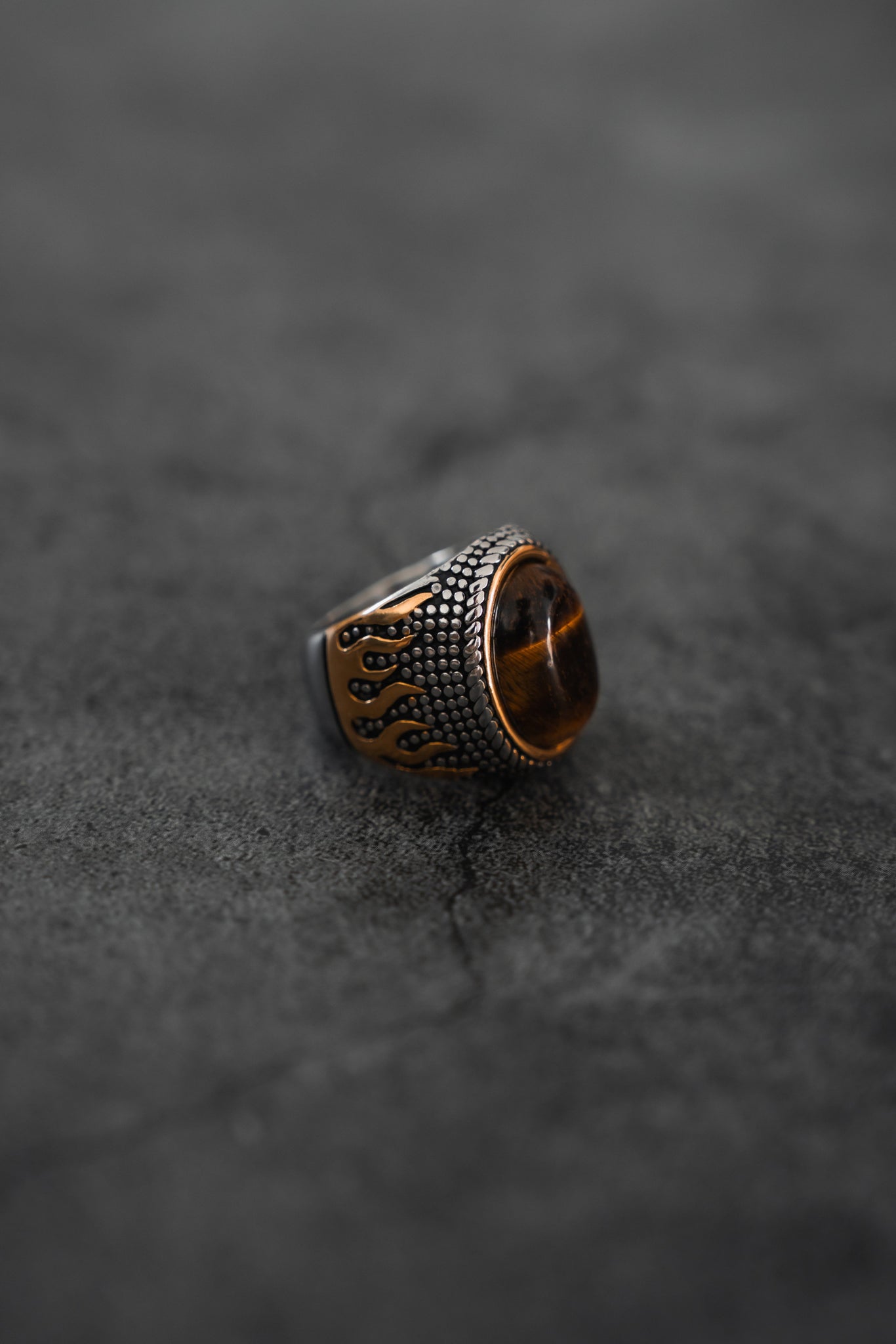 The Relentless Betrayal Inferno Ring features a large brown stone set in a handcrafted, antique-style stainless steel band, displayed close-up on a dark, textured surface.