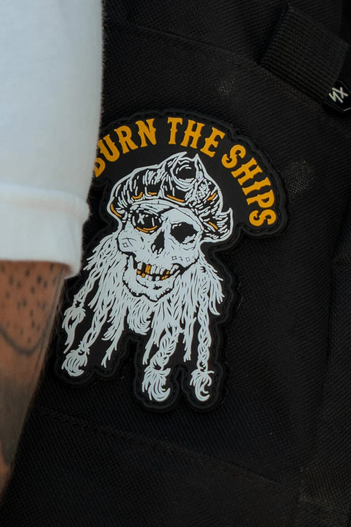 The Burn The Ships PVC Patch by Relentless Betrayal shows a skull with tentacles and the text in yellow, representing creative piracy. Stitched on dark fabric beside a tattooed white sleeve, it symbolizes rebellion and destruction.