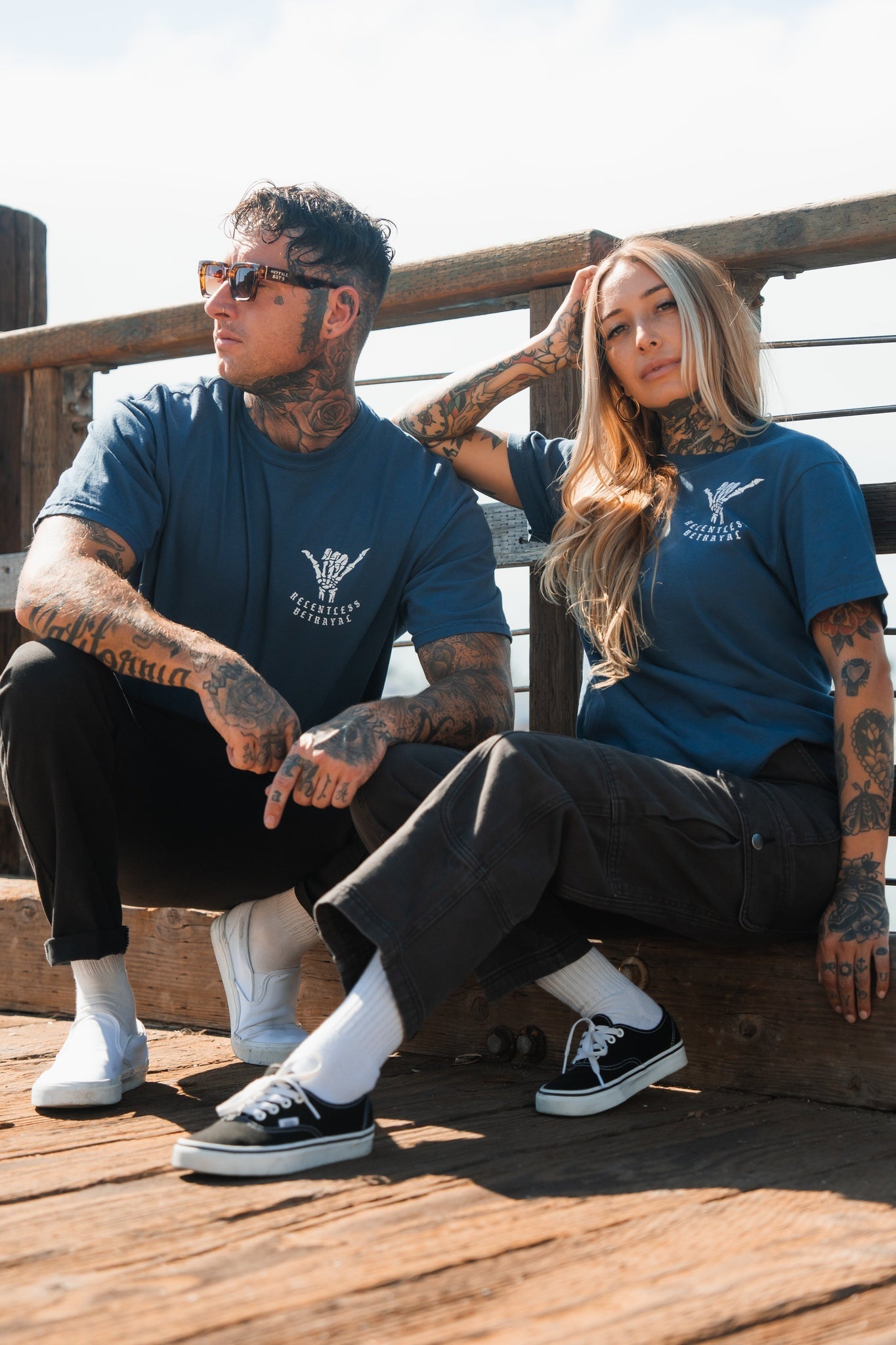 On a wooden deck, two tattooed individuals showcase resilience wearing Sink or Swim Premium Slate T-Shirts from Relentless Betrayal and sunglasses. One confidently sits on a railing while the other squats beside them, with the structure reflecting their inner strength.