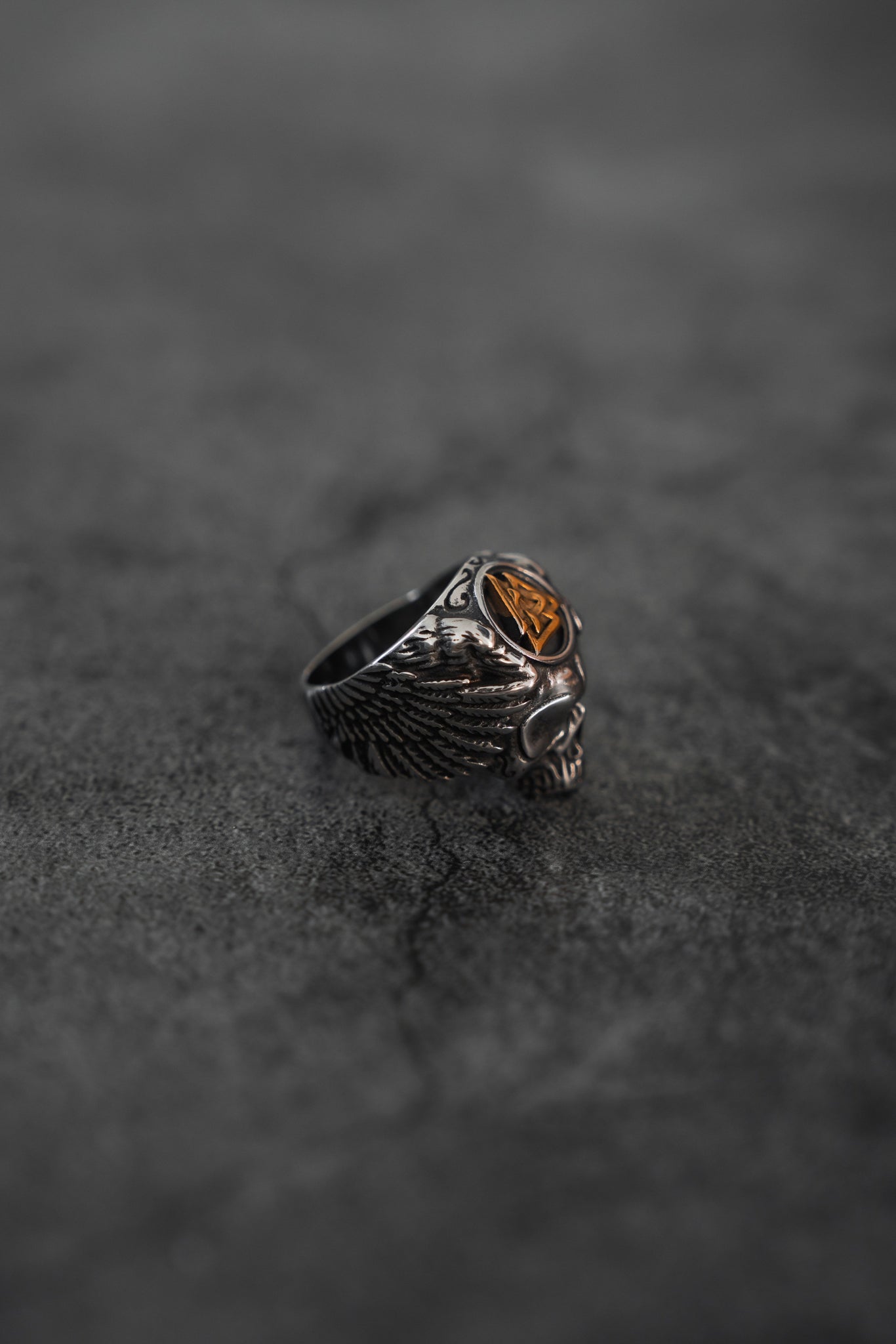 A close-up of the Relentless Betrayals All Gas No Brakes Ring, featuring a detailed skull with wings and a golden shield on stainless steel. The striking contrast against a dark textured surface embodies youth cultures essence.