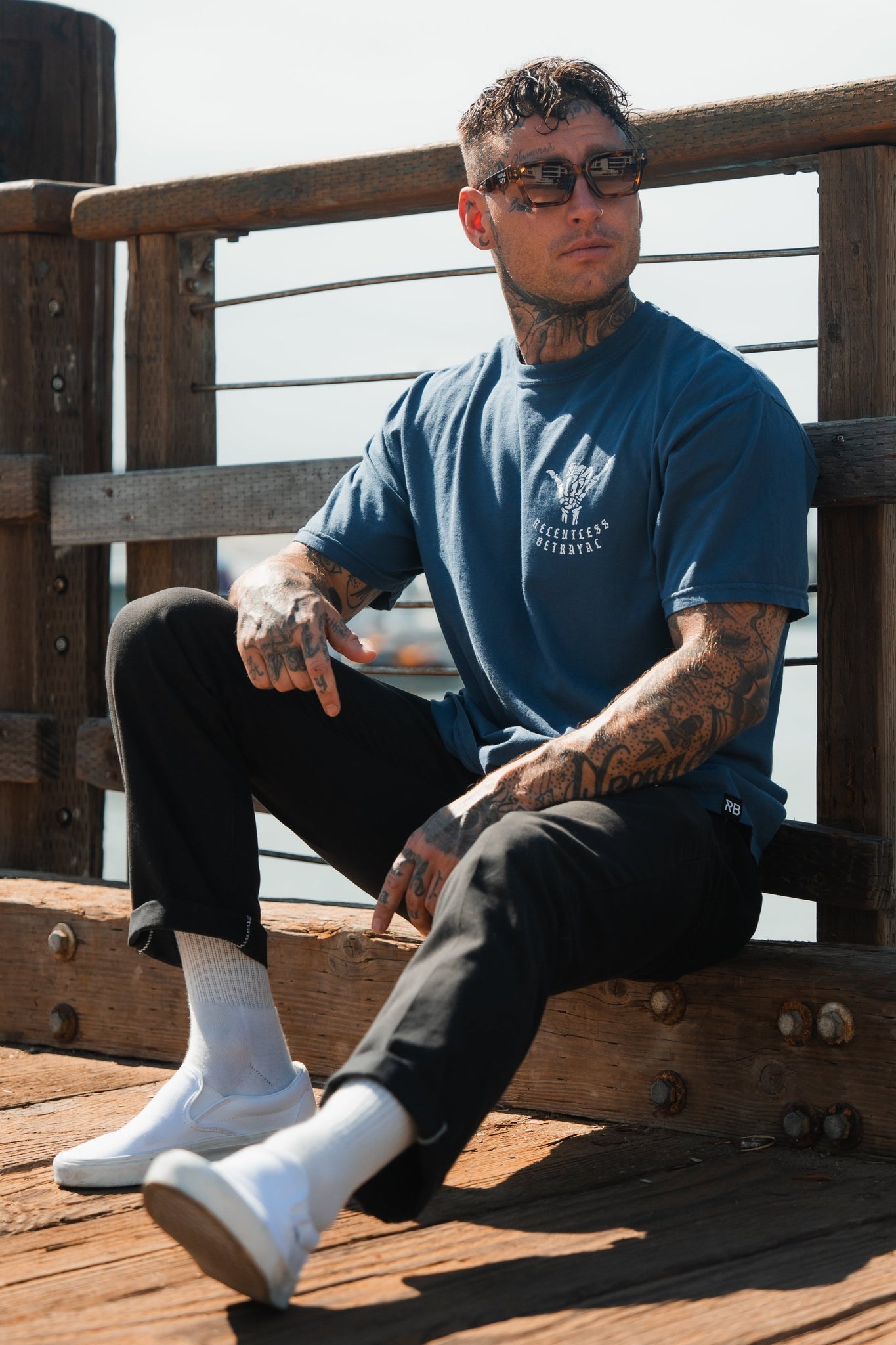 A tattooed man on a wooden deck embodies resilience in his blue “Sink or Swim” Premium Slate T-Shirt by Relentless Betrayal, black pants, white socks, and sneakers. With sunglasses and short hair against a clear sky and wooden railing, he exudes inner strength.