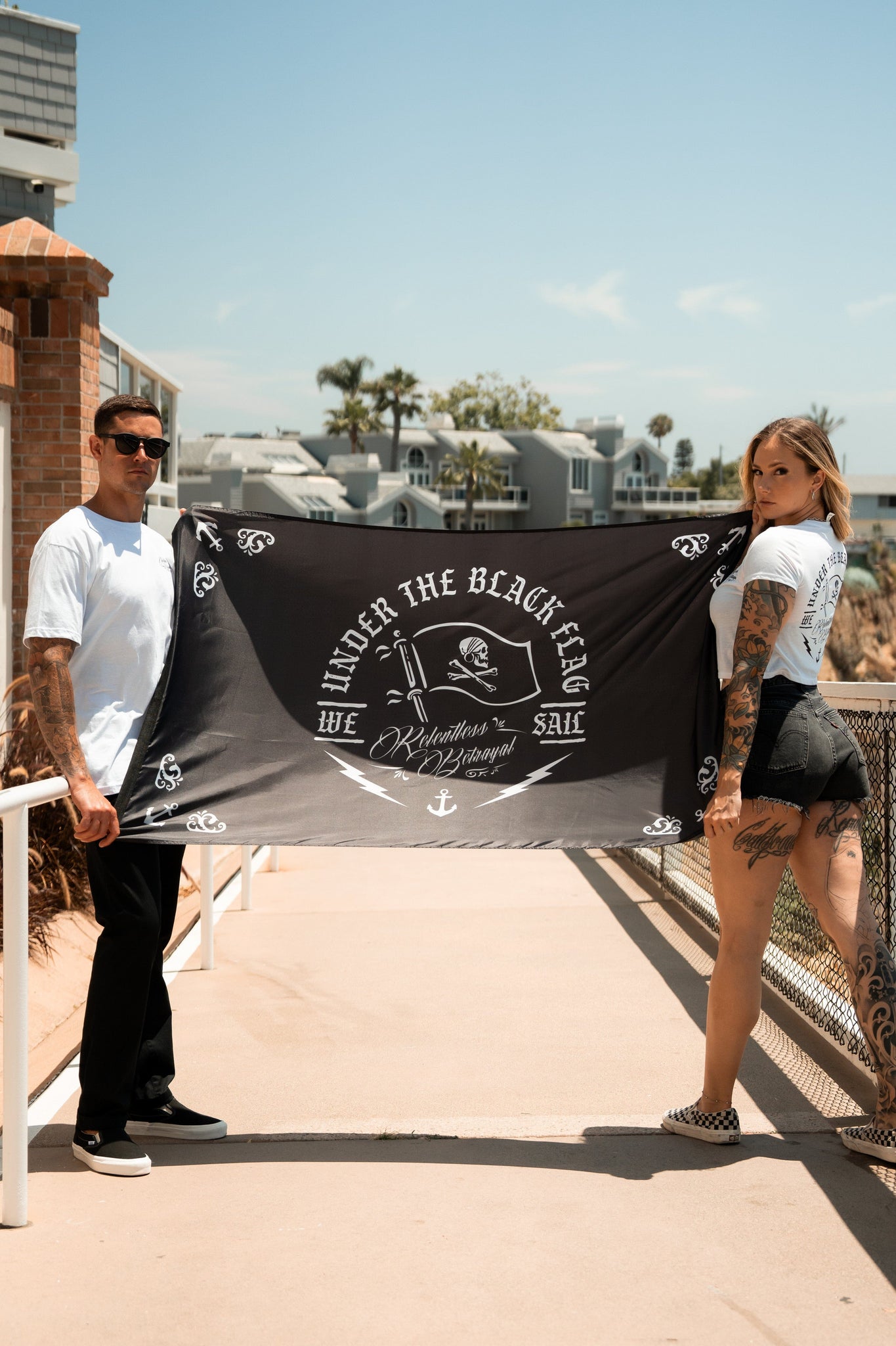 Two people display the Under The Black Flag We Sail flag by Relentless Betrayal, featuring a ship symbol. Crafted from durable polyester with metal grommets, it flutters against a sunny boardwalk backdrop with buildings and palm trees. Both wear casual clothing and have visible tattoos.