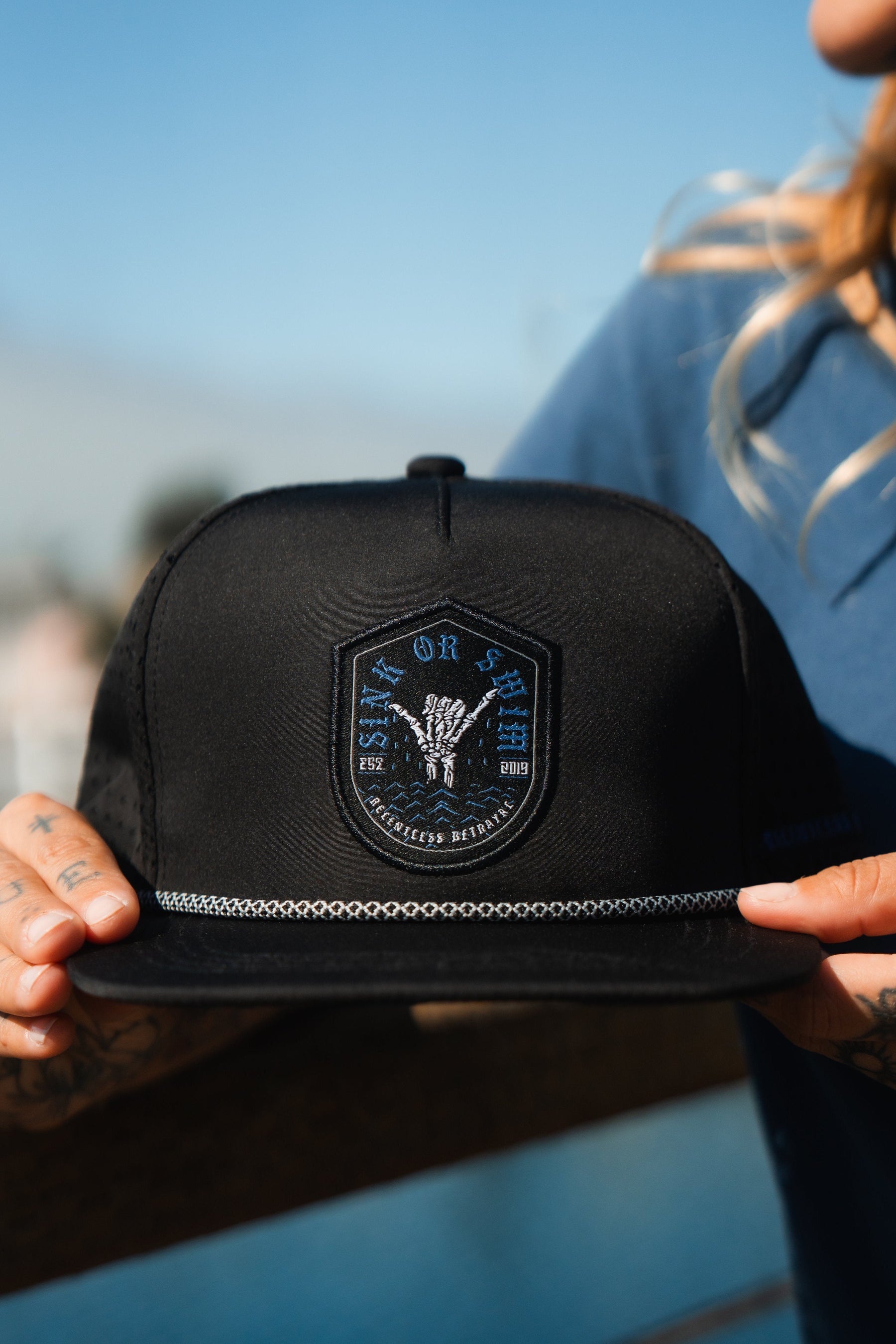 A person confidently holds a Sink Or Swim Snapback from Relentless Betrayal, featuring a patch that reads Live or Die and Restless Dreams, embodying resilience. In the blurred background, their blue shirt emphasizes perseverance in lifes trials.