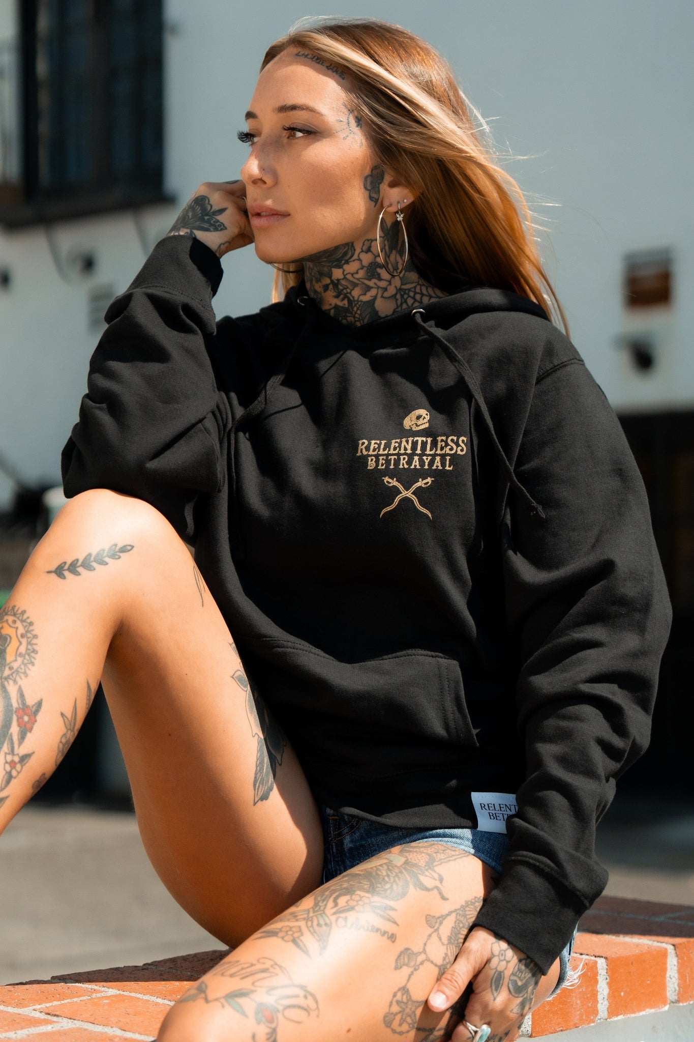 A tattooed individual with long hair, wearing denim shorts and a black cotton/polyester unisex hoodie featuring Loose Lips Sink Ships by Relentless Betrayal, sits outdoors on bricks. They gaze away from the camera with one hand on their chin.