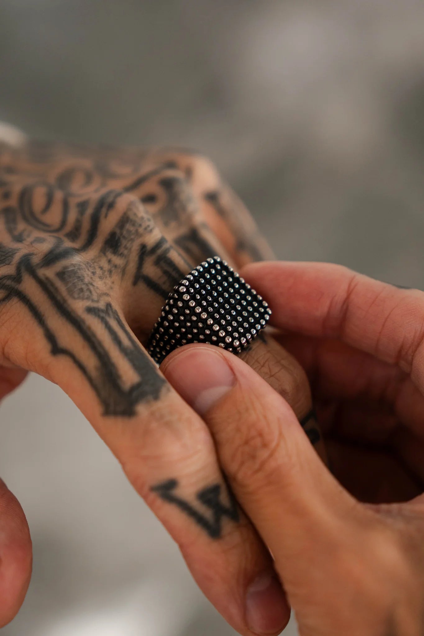 A tattooed hand is seen placing the handcrafted, textured Seeker Ring by Relentless Betrayal on a finger. The intricate tattoo designs adorn the hand, set against a blurred background that highlights both the ring and ink details.
