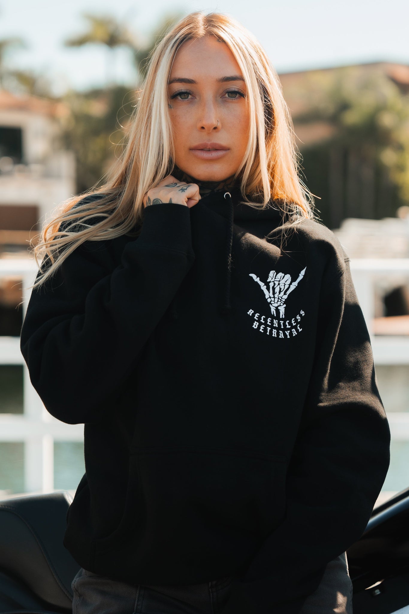 A woman with long blonde hair stands outdoors in a black Sink or Swim Premium Hoodie by Relentless Betrayal, featuring a skeletal hand graphic and resilience-themed text. She grips the collar with her right hand as the sunny, softly blurred background reflects her perseverance.
