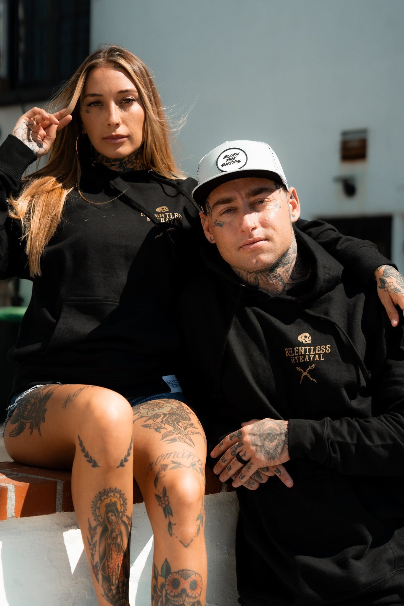 A tattooed man and woman sit outdoors in cozy unisex hoodies. The long-haired woman and the man with a baseball cap wear black Loose Lips Sink Ships Gold Foil Hoodies from Relentless Betrayal, made of a soft cotton/polyester blend, exuding relaxed confidence.