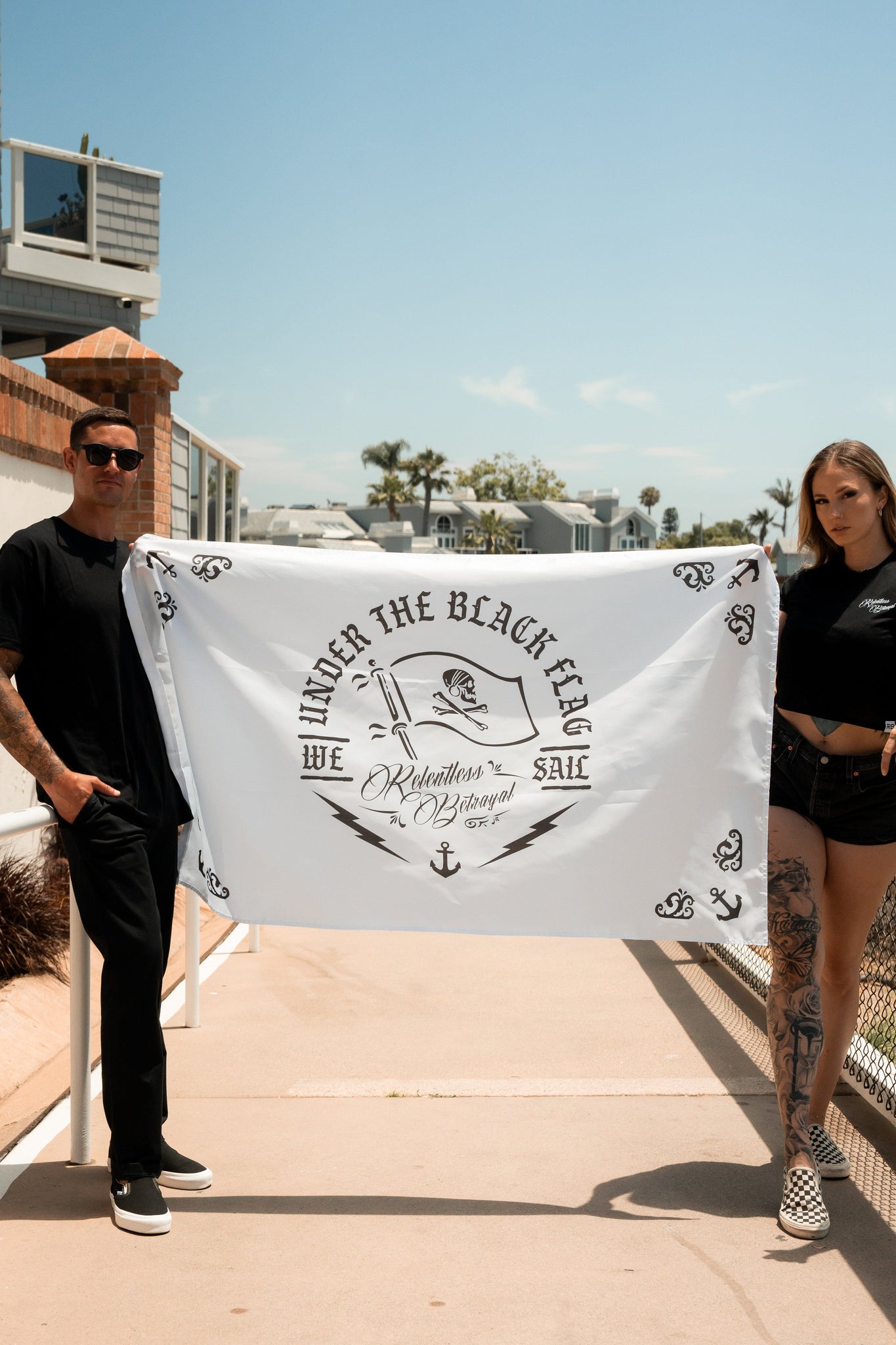 Two people outside hold the Under The Black Flag We Sail V2 polyester flag by Relentless Betrayal, featuring anchors and black text. The person on the left wears sunglasses, and the one on the right has tattooed legs.