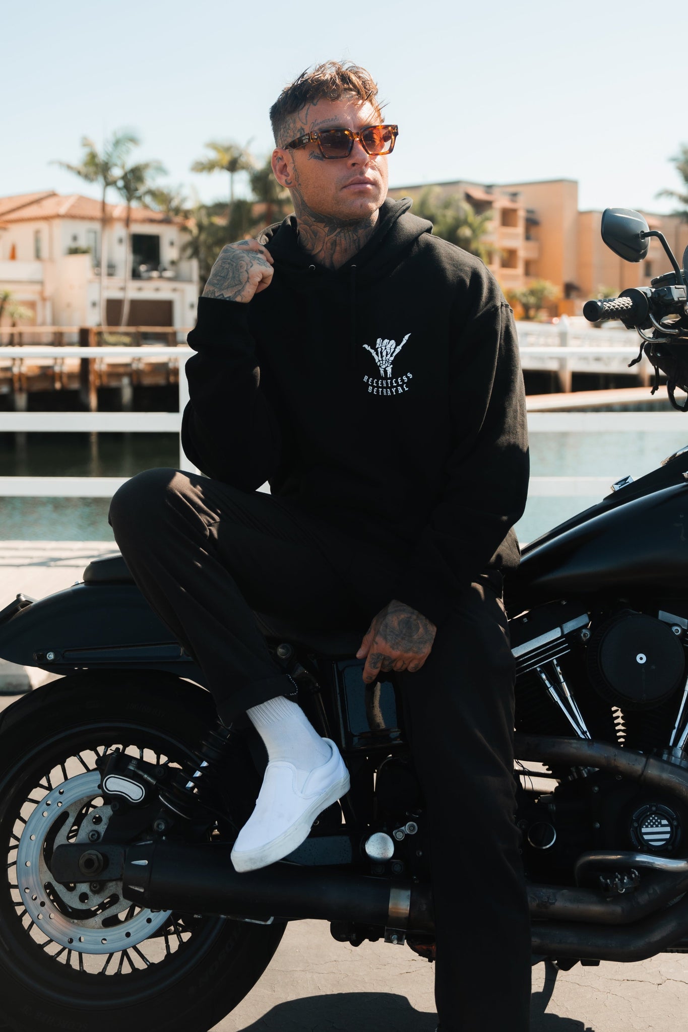 A tattooed person in a black Sink or Swim Premium Hoodie by Relentless Betrayal, wearing sunglasses and white shoes, sits on a motorcycle near the water. They exude resilience with their leg over the bike as buildings and palm trees sway in the backdrop.
