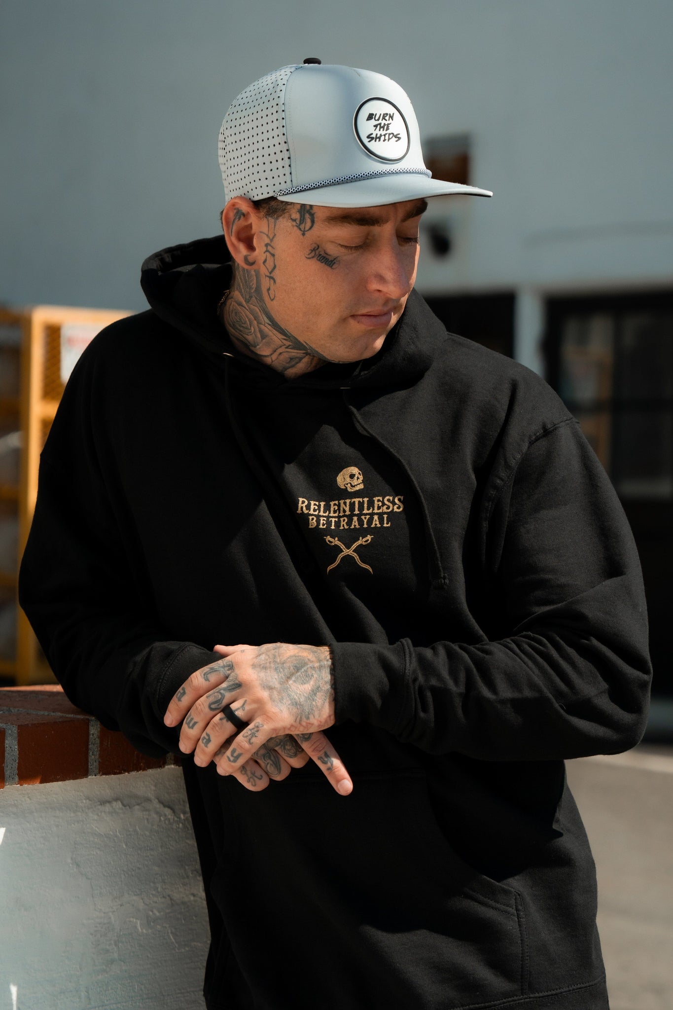 A tattooed individual outdoors leans against a wall, wearing a Relentless Betrayal Loose Lips Sink Ships Gold Foil black cotton/polyester blend hoodie and a white cap. Theyre looking down, hands together, partly in sunlight.