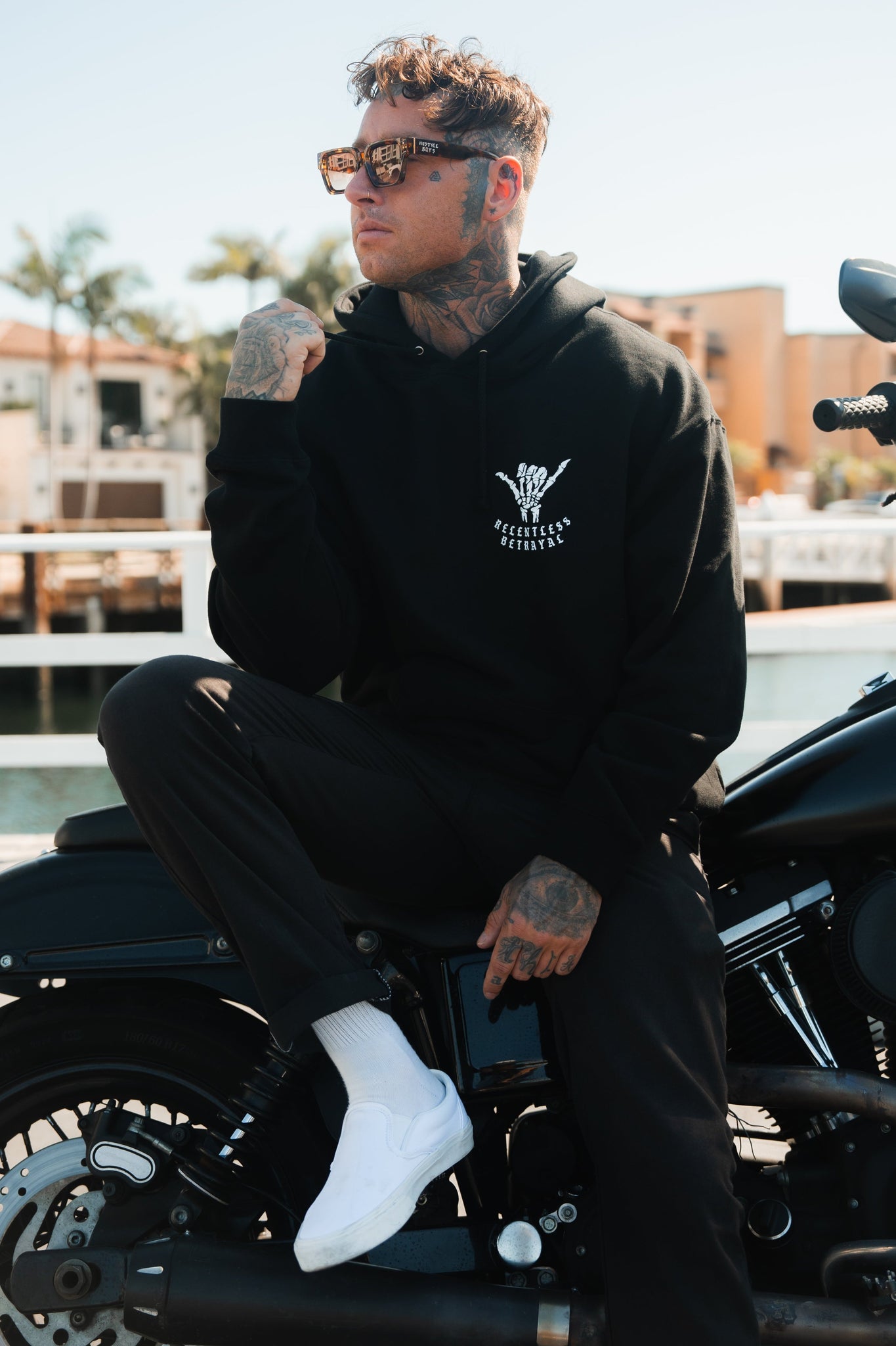 A tattooed person sits confidently on a motorcycle, embodying resilience in a Relentless Betrayal Sink or Swim Premium Hoodie, black pants, and white shoes. One hand near their chin they look to the side, framed by palm trees and buildings.