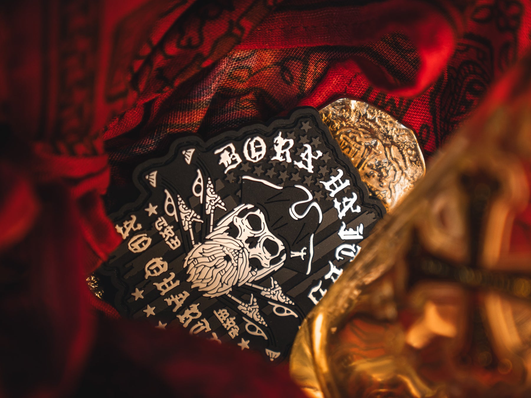 The No Quarter PVC Patch by Born United Private Label displays a skull with Born Through Mercy, set amidst gold details and red fabric. The design includes stylized text and eye symbols against a dark background for a mystical, ornate look.