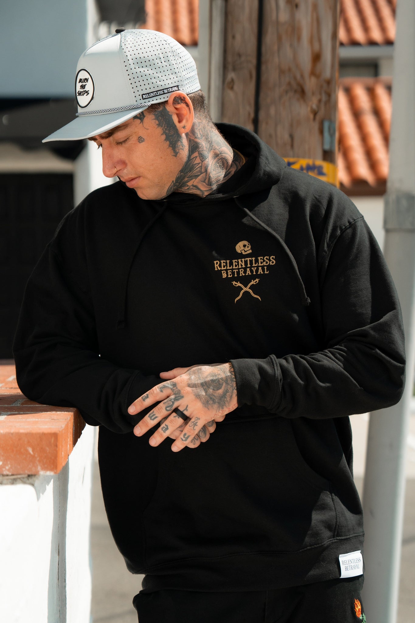 A man in a black Loose Lips Sink Ships Gold Foil hoodie by Relentless Betrayal and white cap leans on a wall outdoors, head tilted down, revealing numerous tattoos. The unisex cotton/polyester blend hoodie complements the tiled roof and wooden post backdrop.