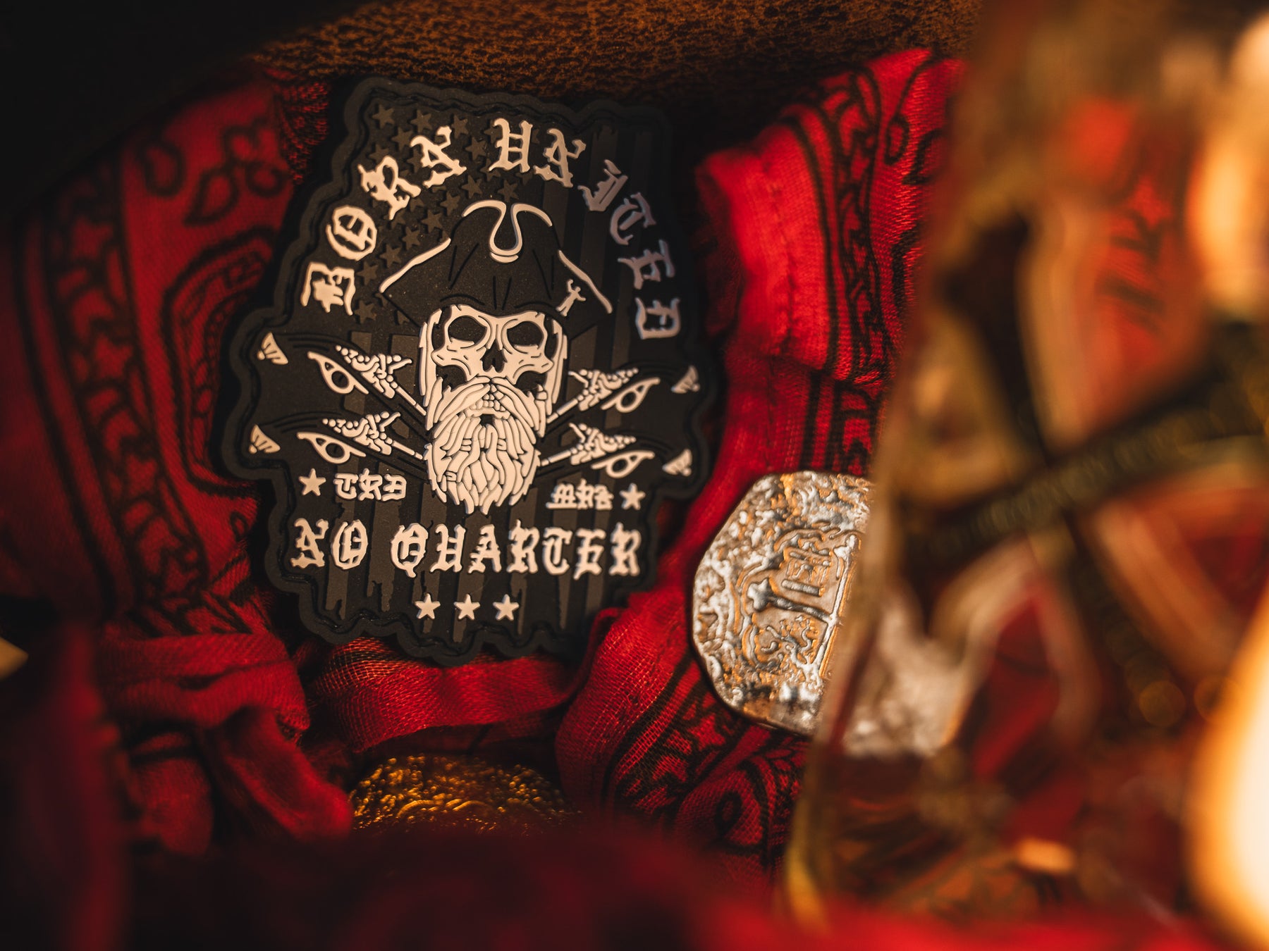 Amid red fabric and shimmering coins, the No Quarter PVC Patch from Born United Private Label features a skull with a pirate hat and crossed swords, boldly declaring Born Unto No Quarter, evoking an aura of hidden treasure in the dimly lit scene.
