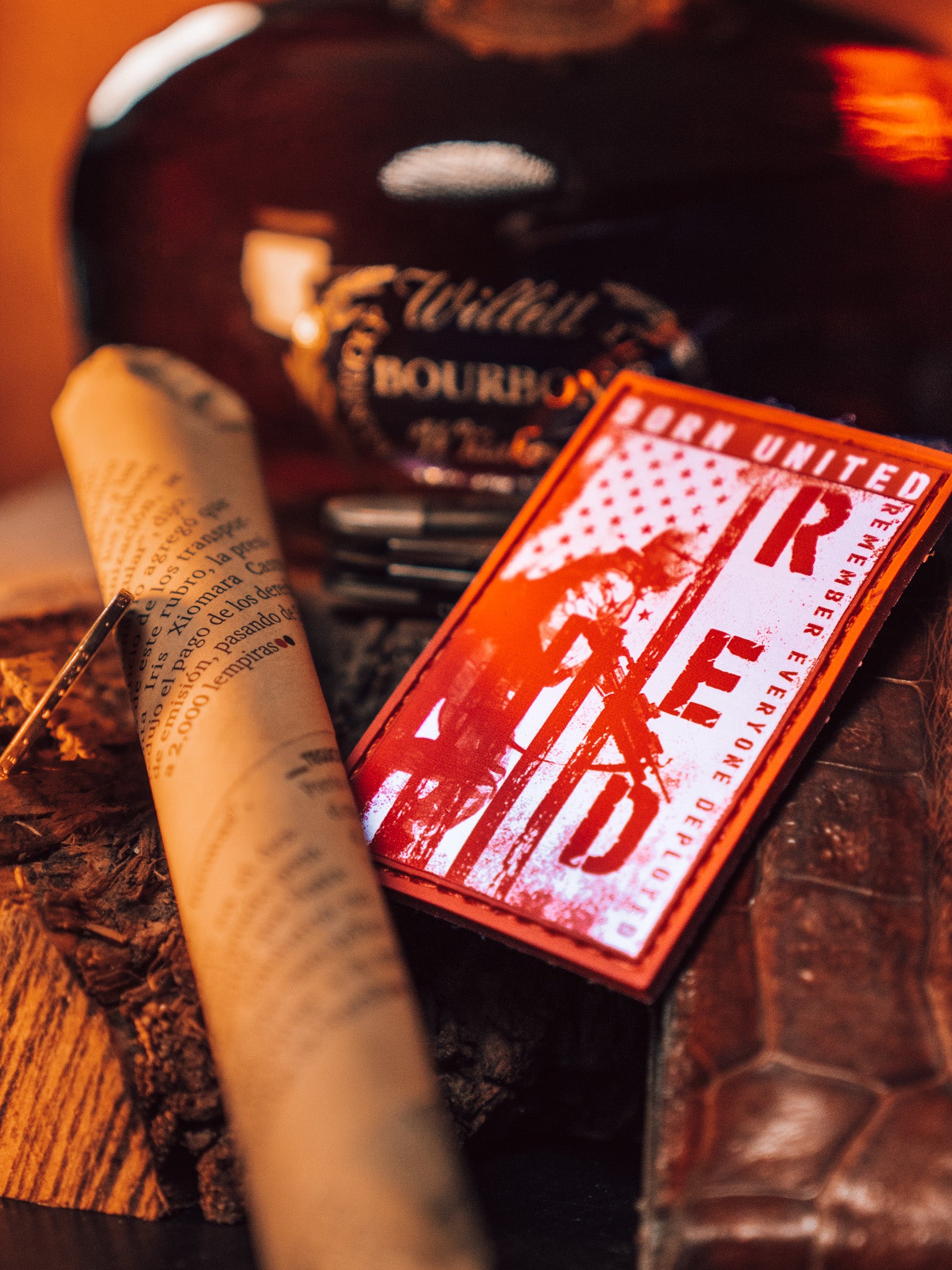 A close-up of the Born United Private Label R.E.D. Friday PVC Patch, featuring the American flag and a soldier silhouette to symbolize military support. Its set against a rolled parchment, brown leather accessory, pen, and bourbon bottle, all in warm lighting to honor Remember Everyone Deployed.
