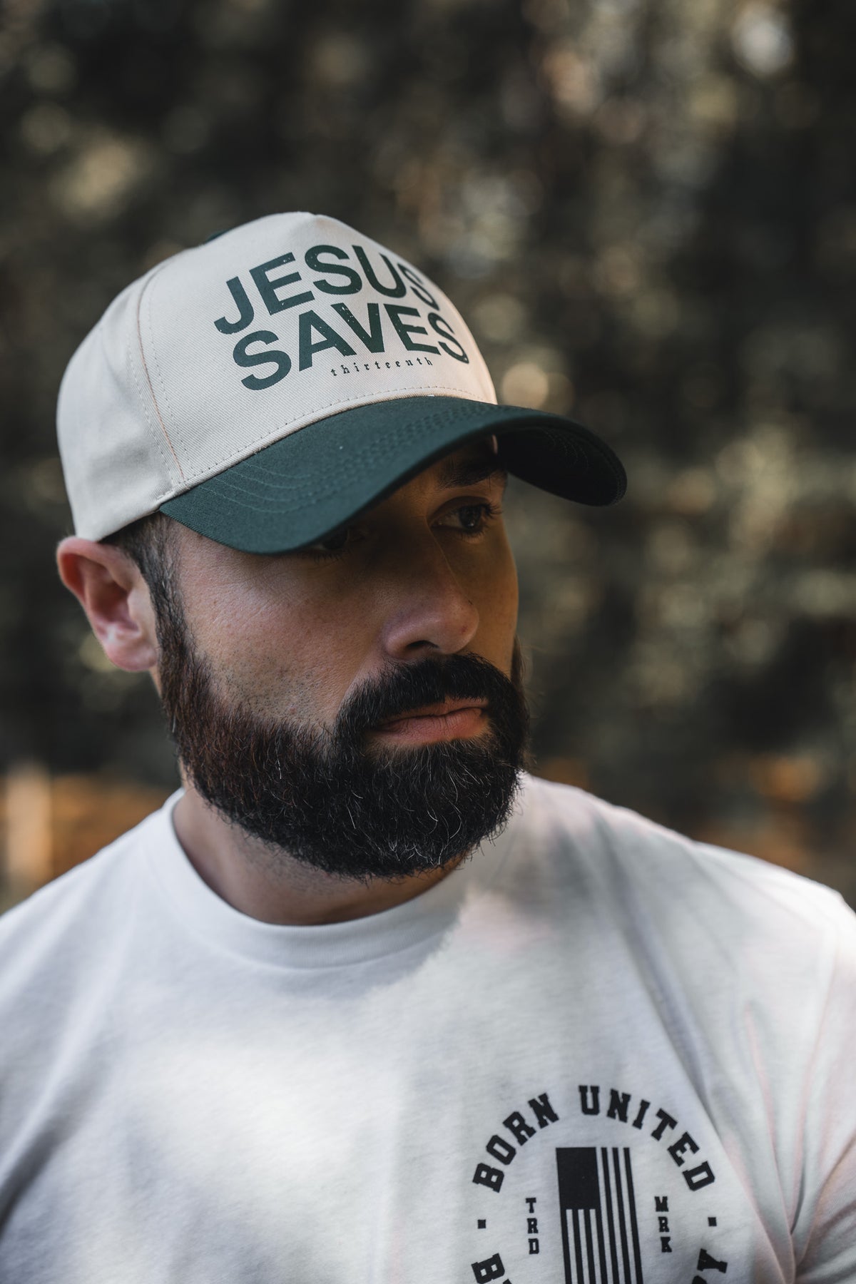 A bearded man wears a 13th Jesus Saves Ivory Snapback and a white tee with a circular BORN UNITED logo, standing outdoors against a blurred natural background that reflects the message of John 3:16.