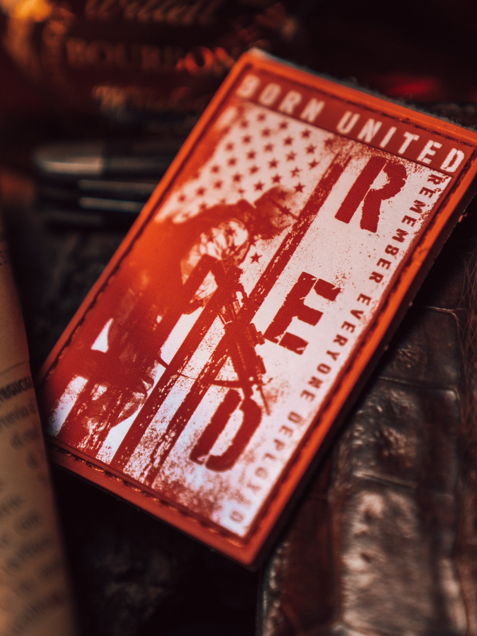 The R.E.D. Friday PVC Patch by Born United Private Label features a striking red and black design with R.E.D and Remember Everyone Deployed text, promoting military support. It includes a stylized American flag and soldier figure, surrounded by dark, blurred objects.