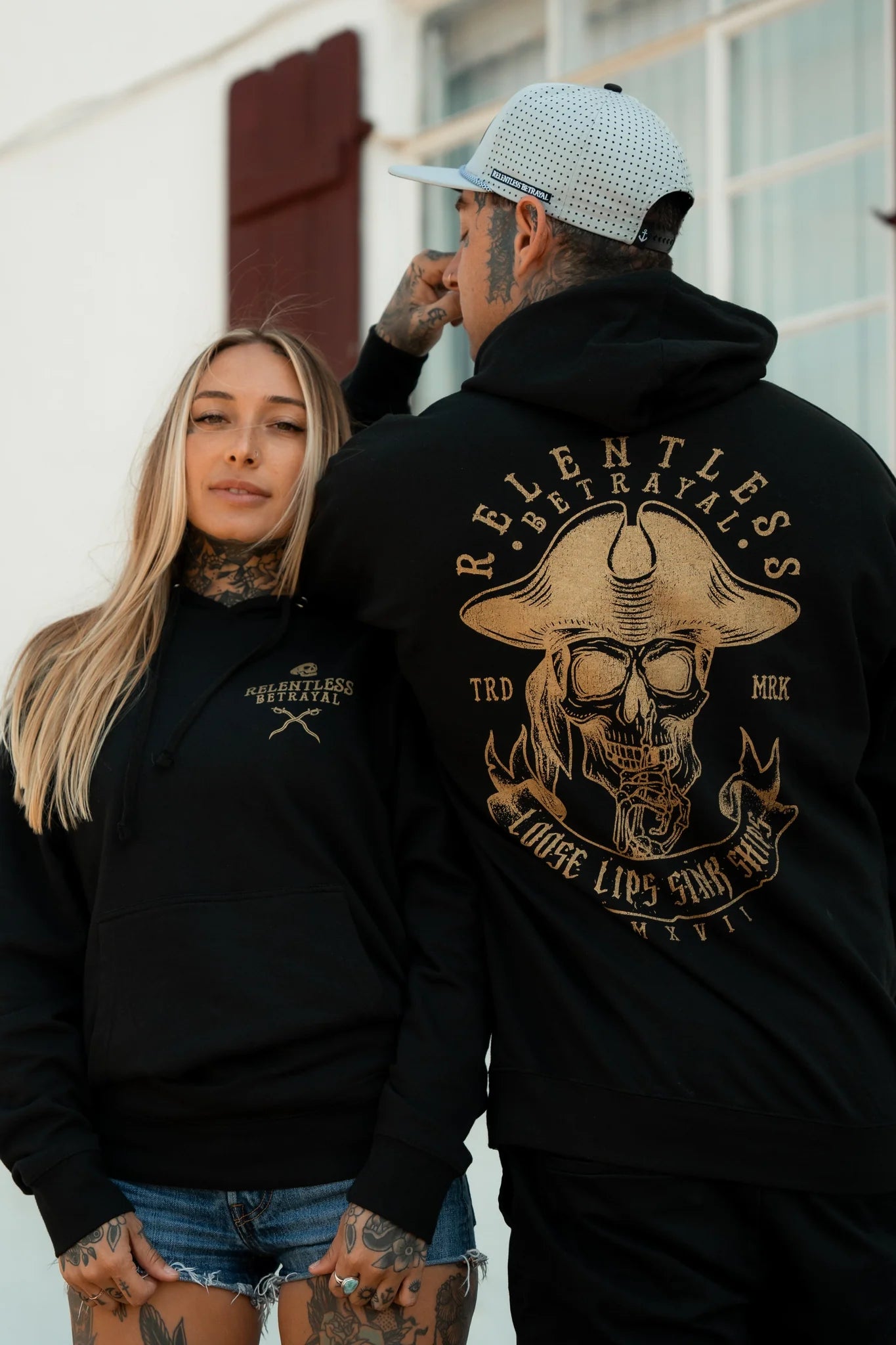 A woman and a man pose confidently, both in unisex hoodies with unique designs. The mans Loose Lips Sink Ships Gold Foil Hoodie from Relentless Betrayal features a large skull graphic on the back. Both are tattooed, standing in front of a white building with a window.