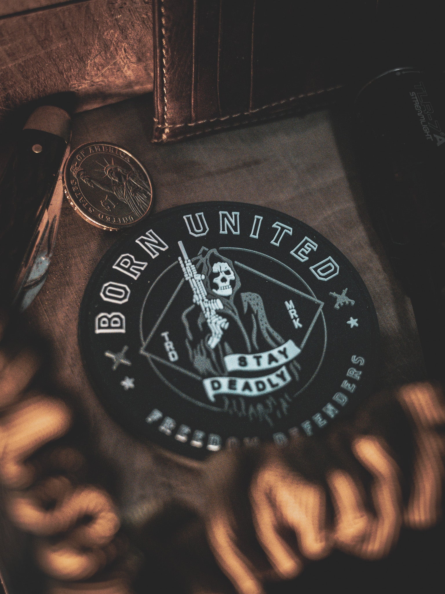 Close-up of the Stay Deadly PVC Patch by Born United Private Label, showcasing a skull with rifle and Born United, Stay Deadly, Freedom Defenders text. Surrounding items include a knife, coin, and wallet on a textured surface under dim dramatic lighting.