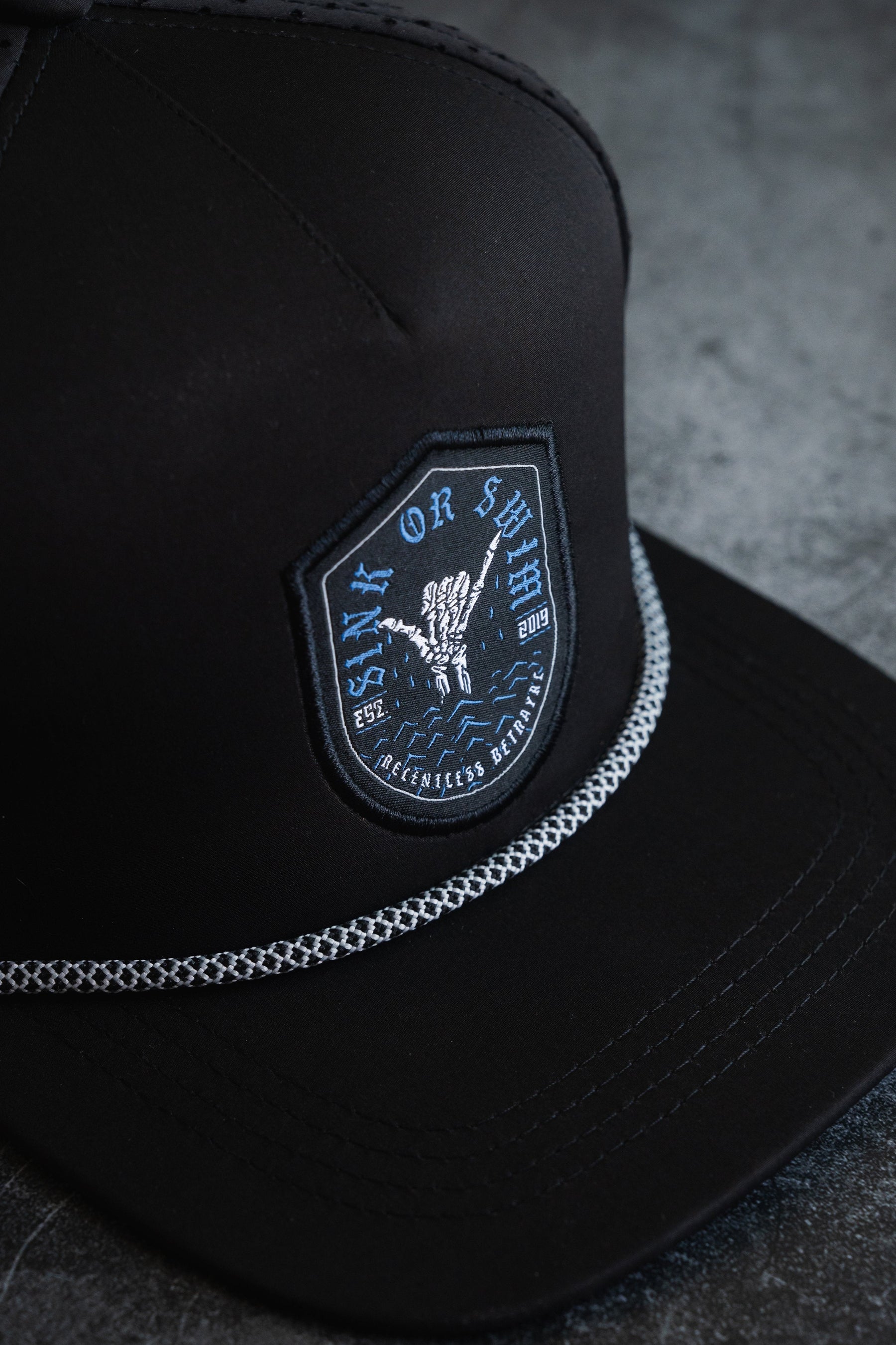 The Sink Or Swim Snapback by Relentless Betrayal features a black cap with an anchor and rope patch, symbolizing resilience. The patch reads Sink or Swim and showcases the years 1922 and 2019 in blue and white on a textured trim.