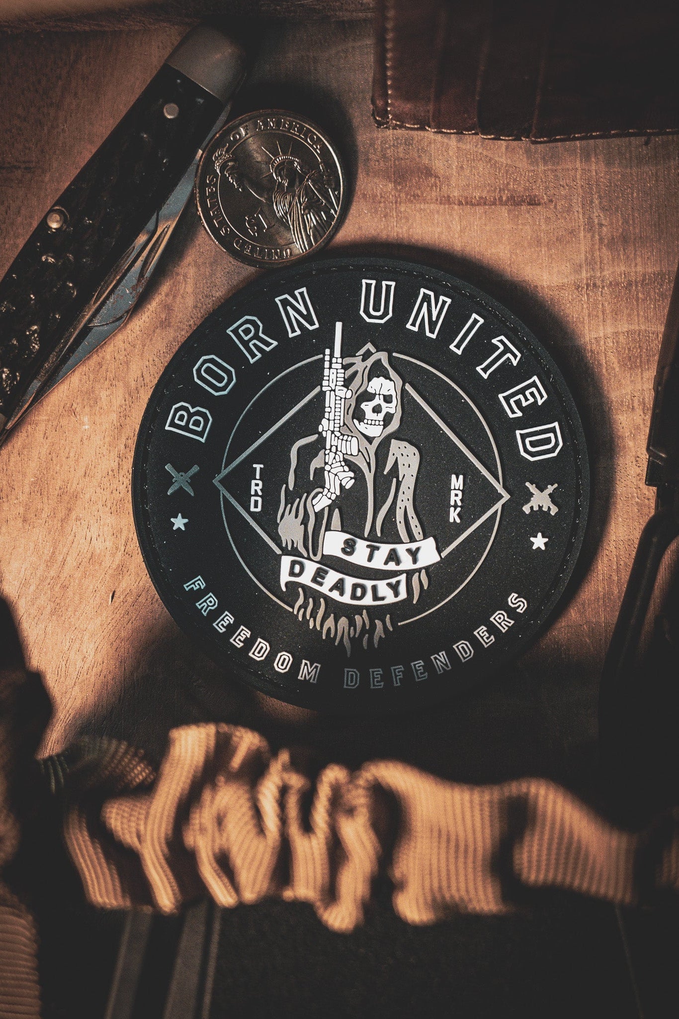 The Stay Deadly PVC Patch by Born United Private Label, featuring a skeleton with a gun and the text Born United, Stay Deadly, and Freedom Defenders, is displayed on wood with a pocket knife and coin.