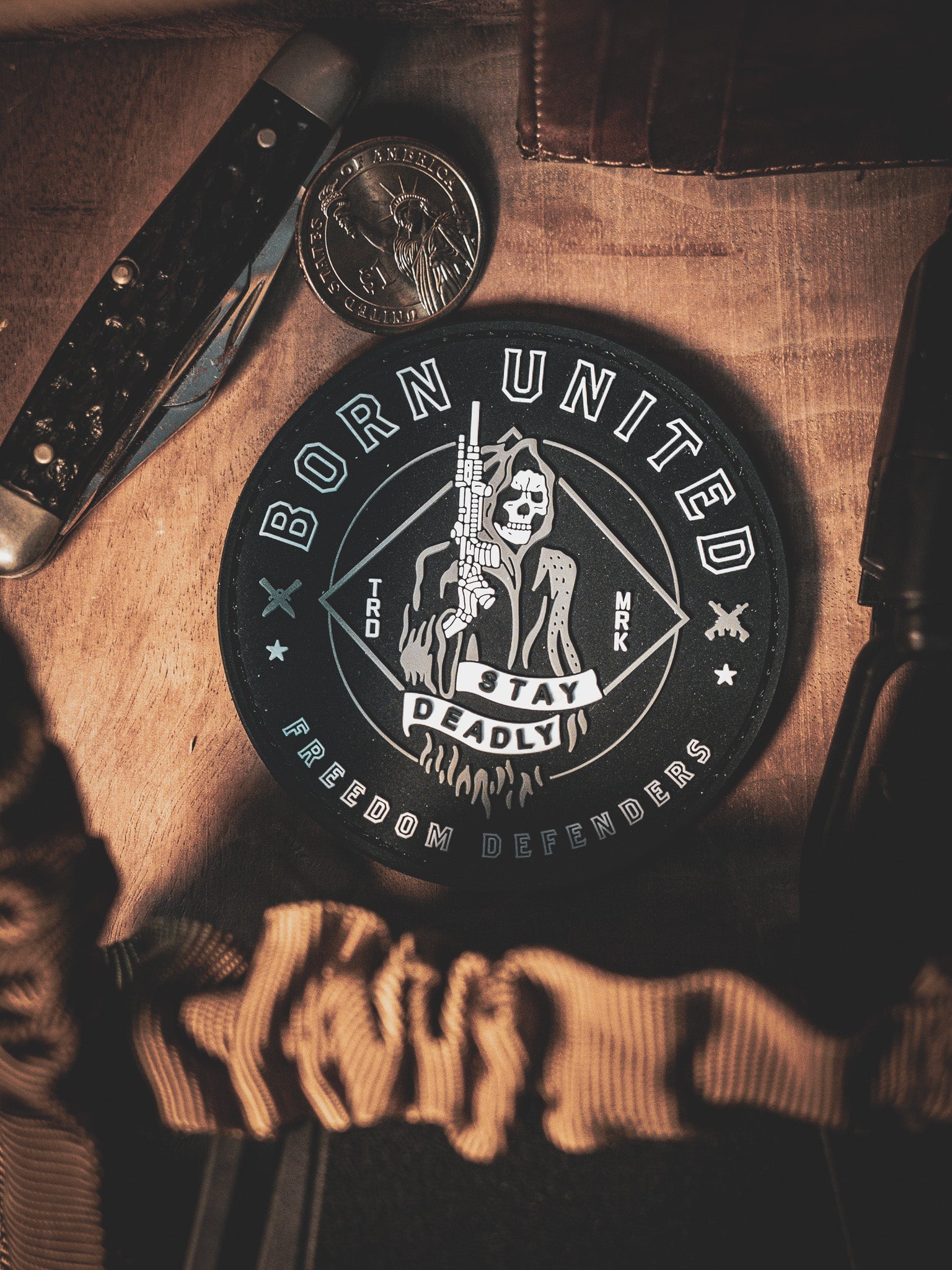 The Stay Deadly PVC Patch by Born United Private Label, featuring a skeleton with a gun and the text Born United, Stay Deadly, and Freedom Defenders, is displayed on wood with a pocket knife and coin.