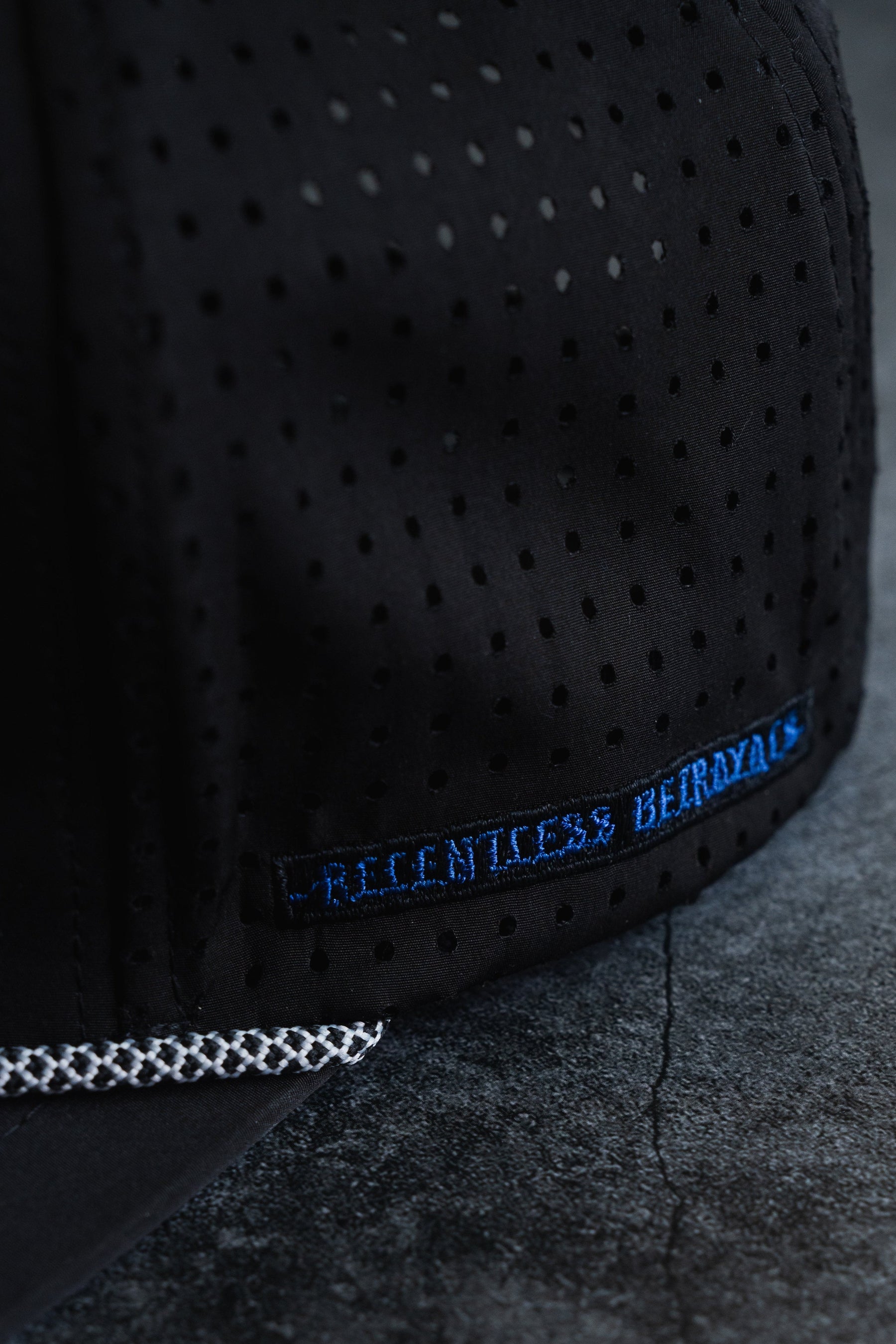 Close-up of a Sink Or Swim Snapback by Relentless Betrayal, featuring a black perforated design and a blue embroidered phrase on the side symbolizing resilience. The cap is placed on a dark, textured surface.