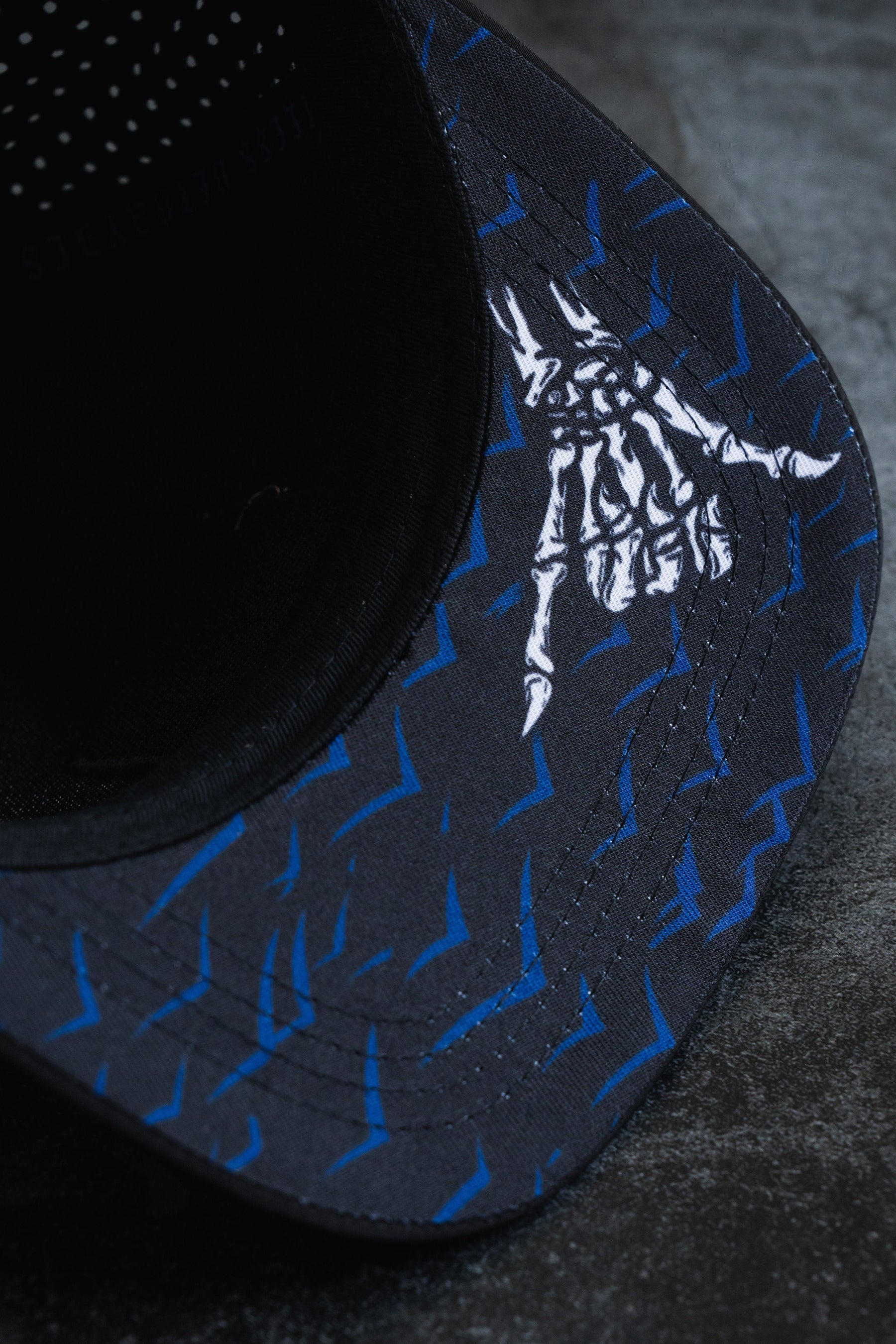 Close-up of a dark Sink Or Swim Snapback by Relentless Betrayal, featuring a blue arrow pattern symbolizing resilience and a white skeleton hand signifying perseverance. The textured background underscores overcoming challenges with style.