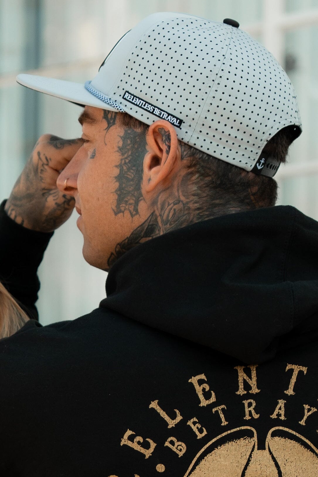 A tattooed individual wears a white Burn The Ships Cement PVC Snapback and a black hoodie with RELENTLESS BETRAYAL on the back, posing outdoors against a sleek glass building.