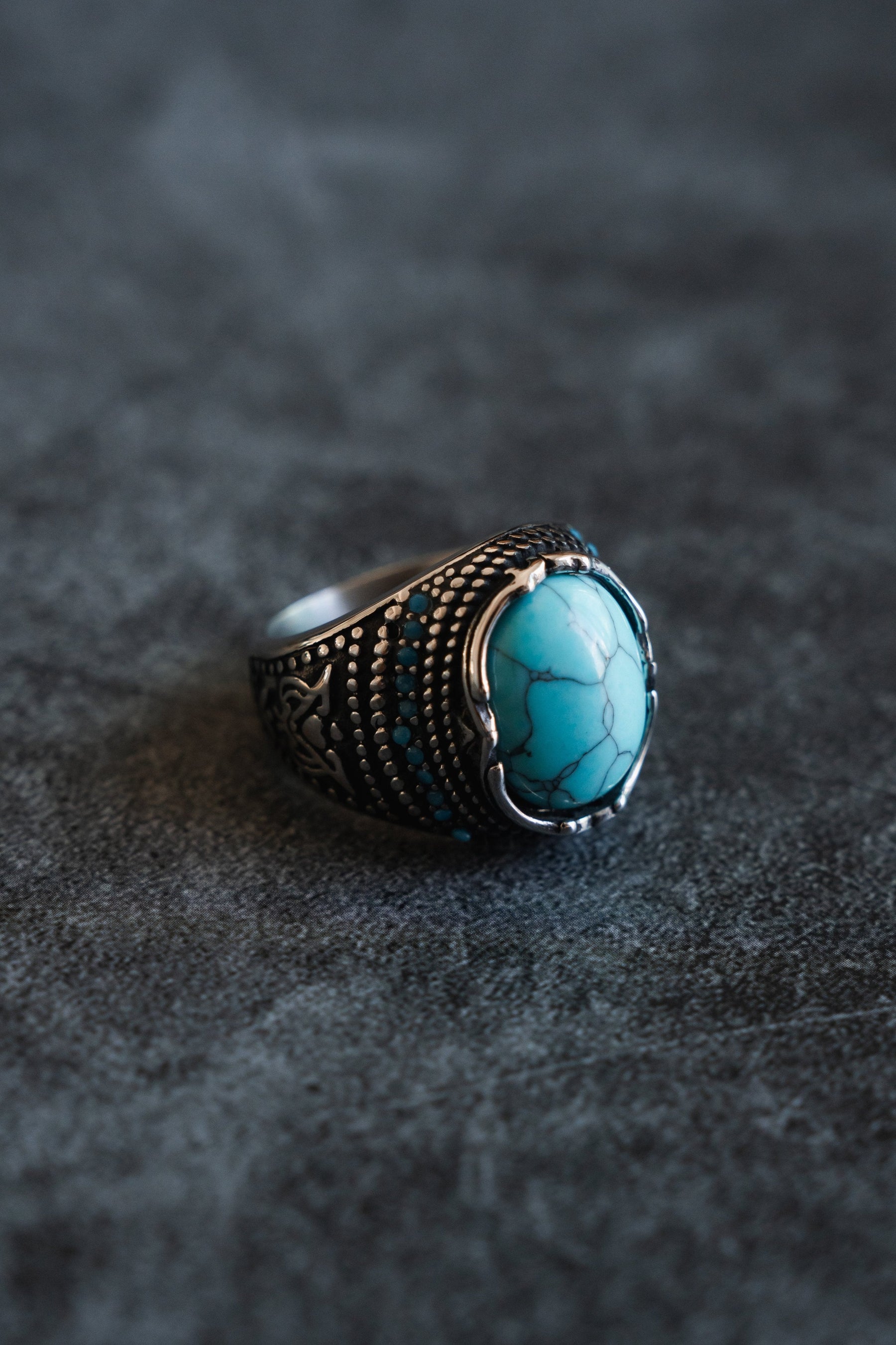 The Eye of the Storm Ring by Relentless Betrayal showcases a handcrafted design with an intricate pattern and a striking oval turquoise stone, elegantly set on a textured, dark surface.