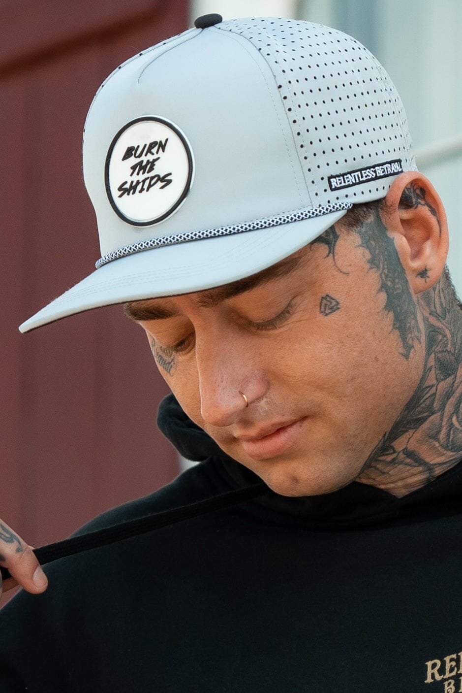A man with tattoos on his neck and face stands in a black hoodie, wearing a perforated white, water-resistant Relentless Betrayal snapback named Burn The Ships Cement PVC, evoking the pirate lifestyle. He gazes downward against a blurred backdrop.