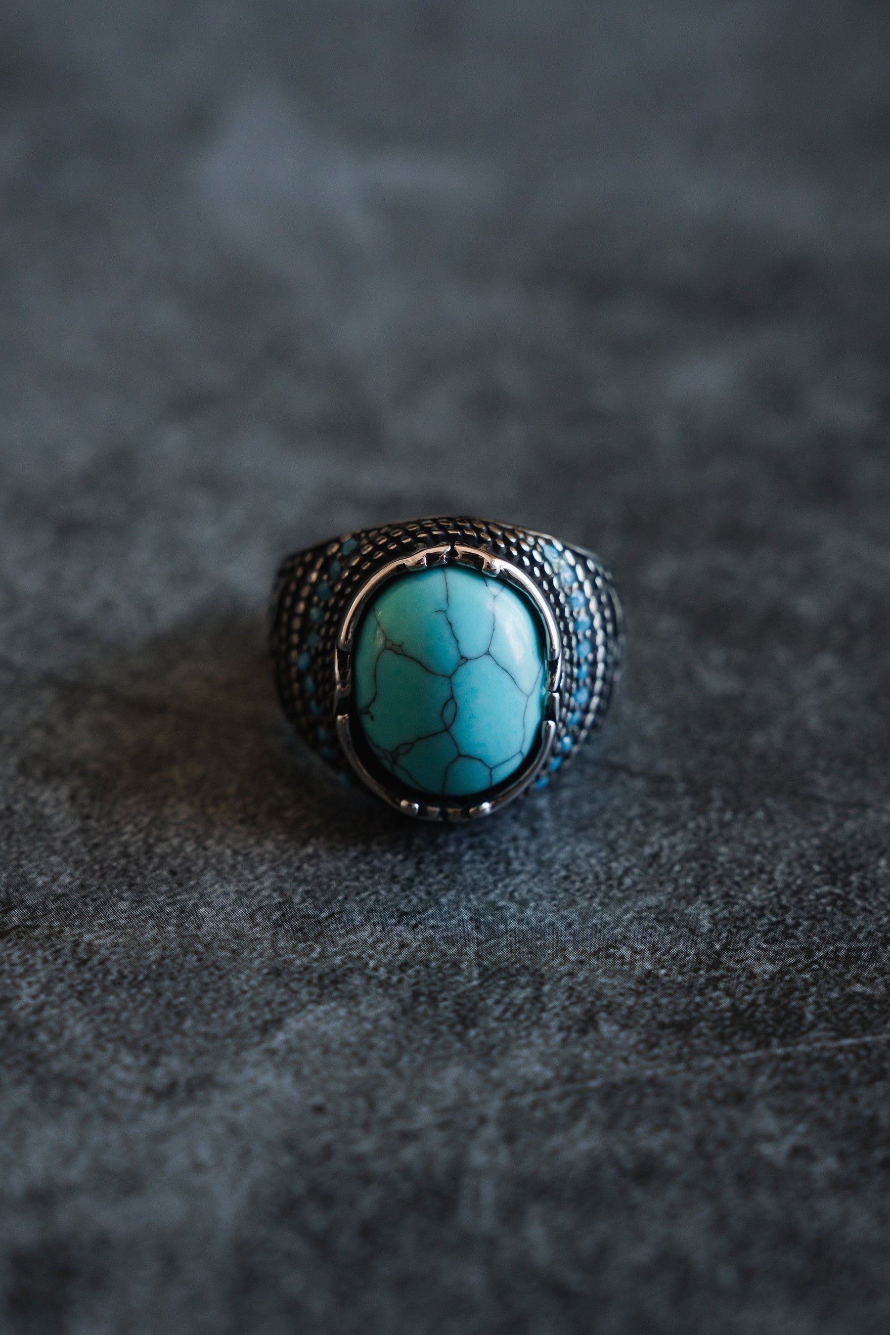 Discover Relentless Betrayals Eye of the Storm Ring, a handcrafted piece featuring a large turquoise gemstone with black veining on a dark textured background, complemented beautifully by an ornate silver band design.
